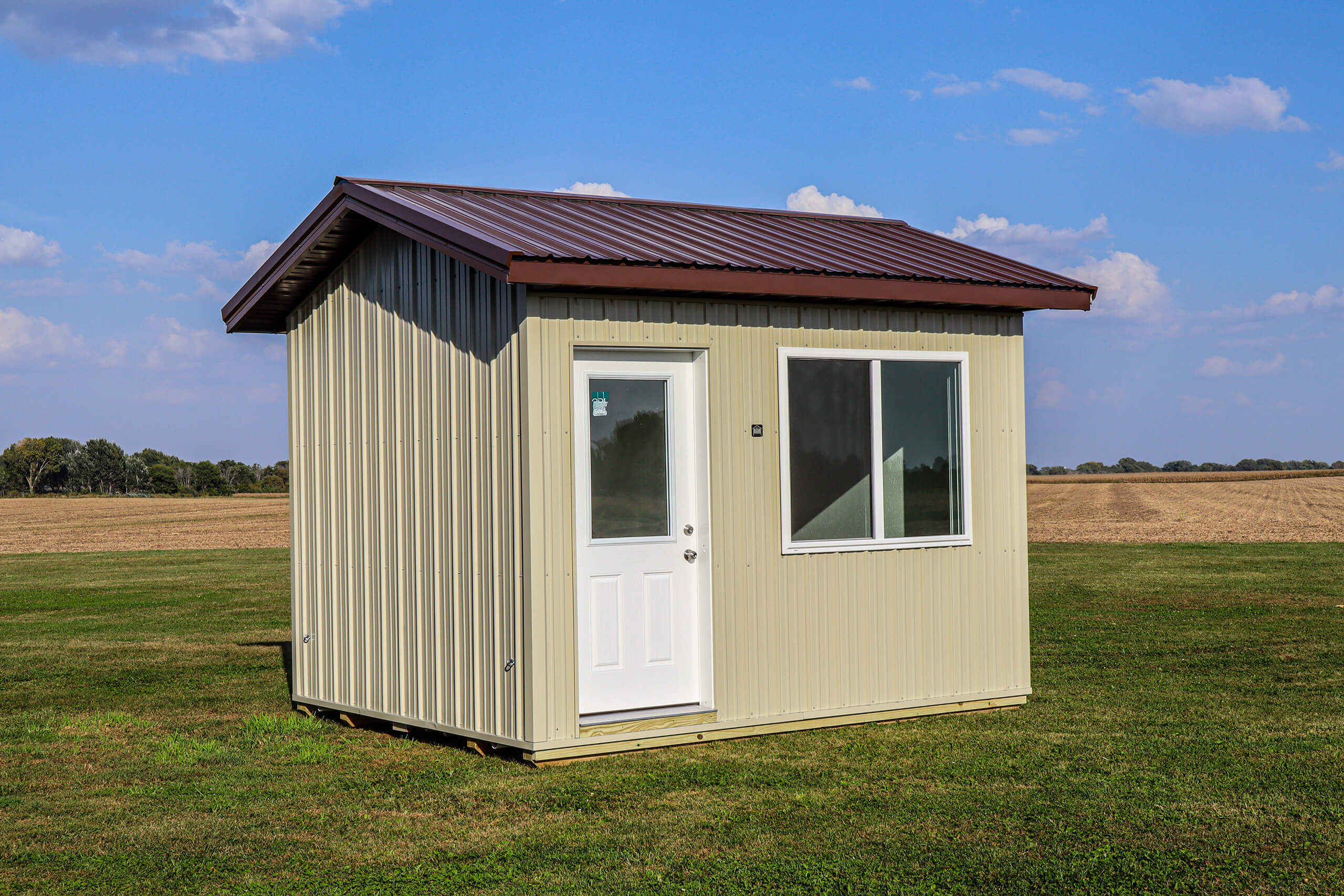 A small cream-colored shed with a metal roof, one door, and two windows, sits on green grass in an open rural area under a blue sky.