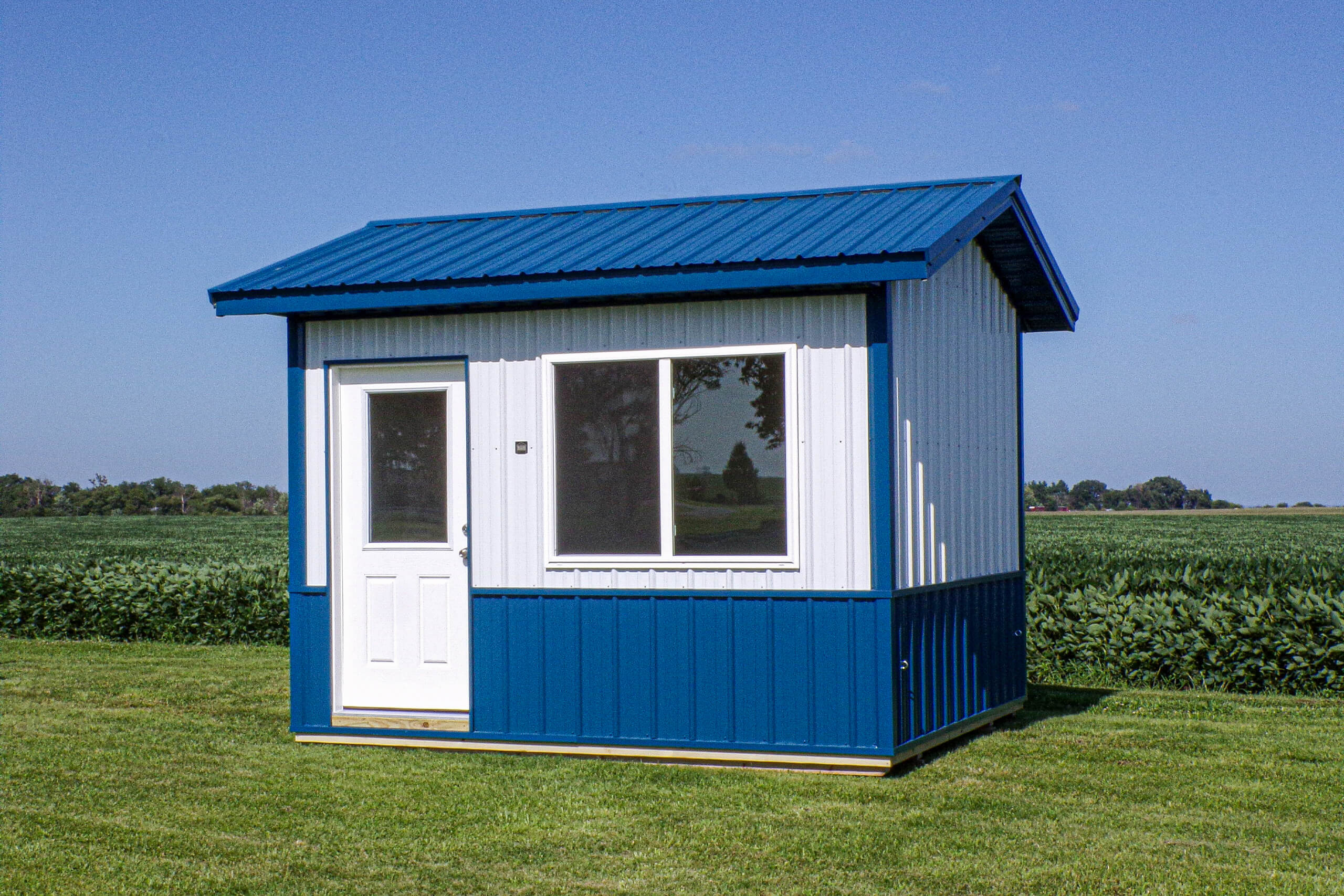 A small blue and white shed with a metal roof, a door, and two windows sits on a grassy field with crops and trees in the background under a clear sky.