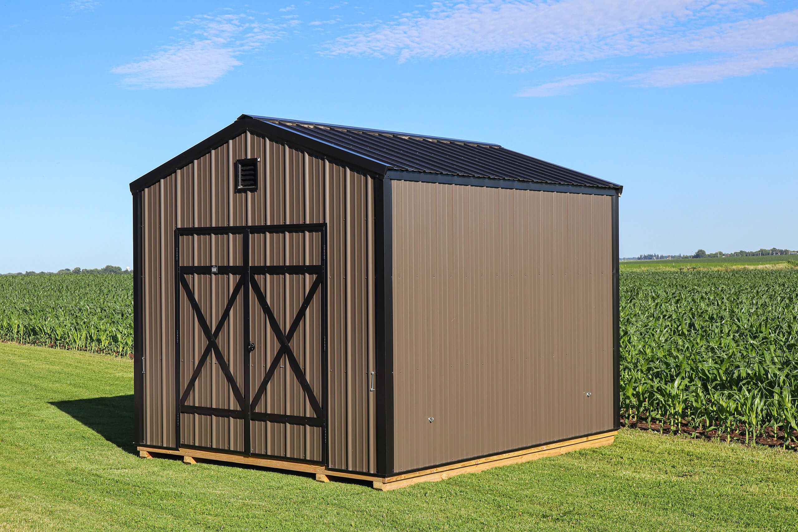 A brown metal storage shed with black trim sits on a grassy field near a crop of young corn under a blue sky with scattered clouds.