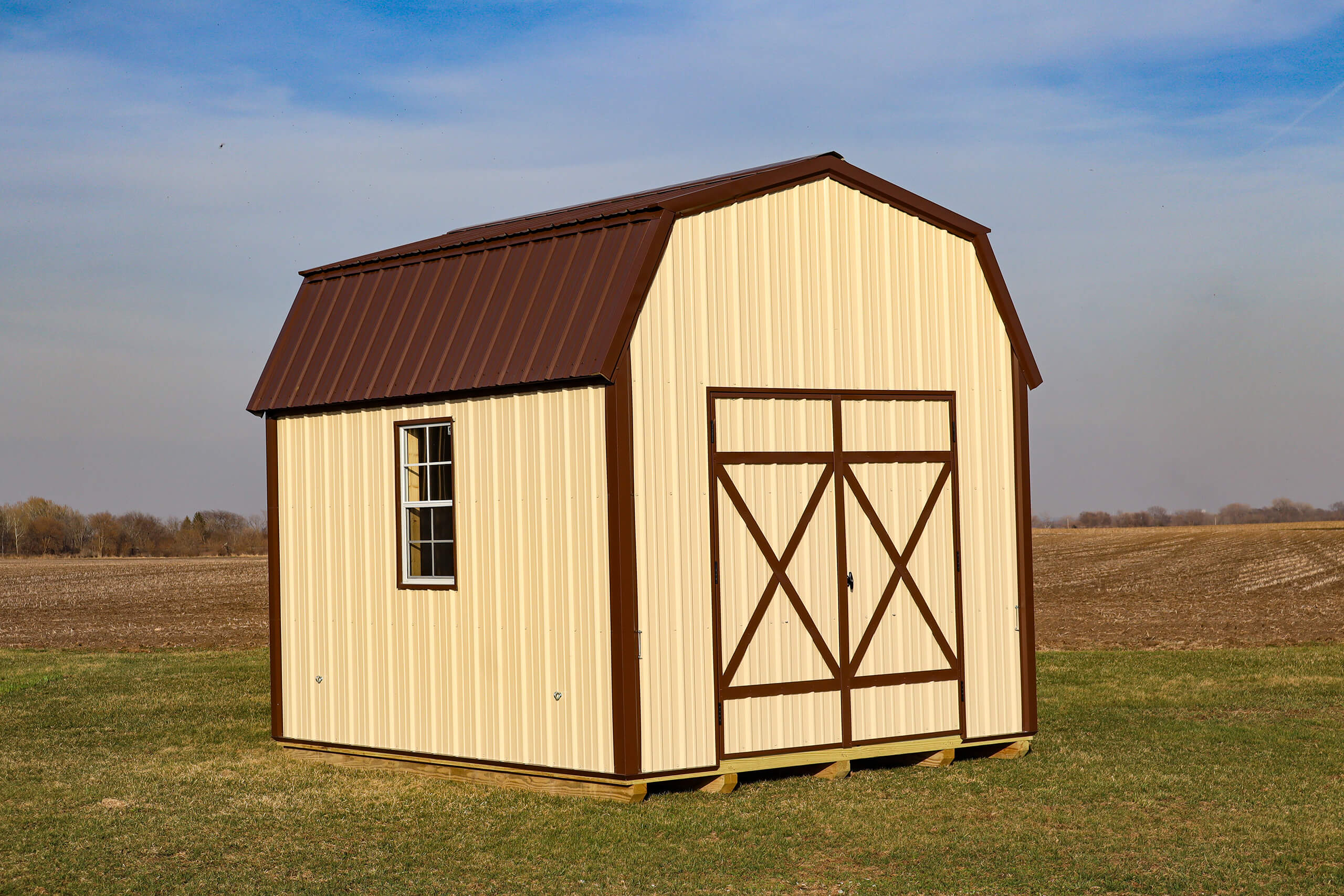 A cream-colored shed with brown trim and a barn-style roof stands on a grassy field under a blue sky.