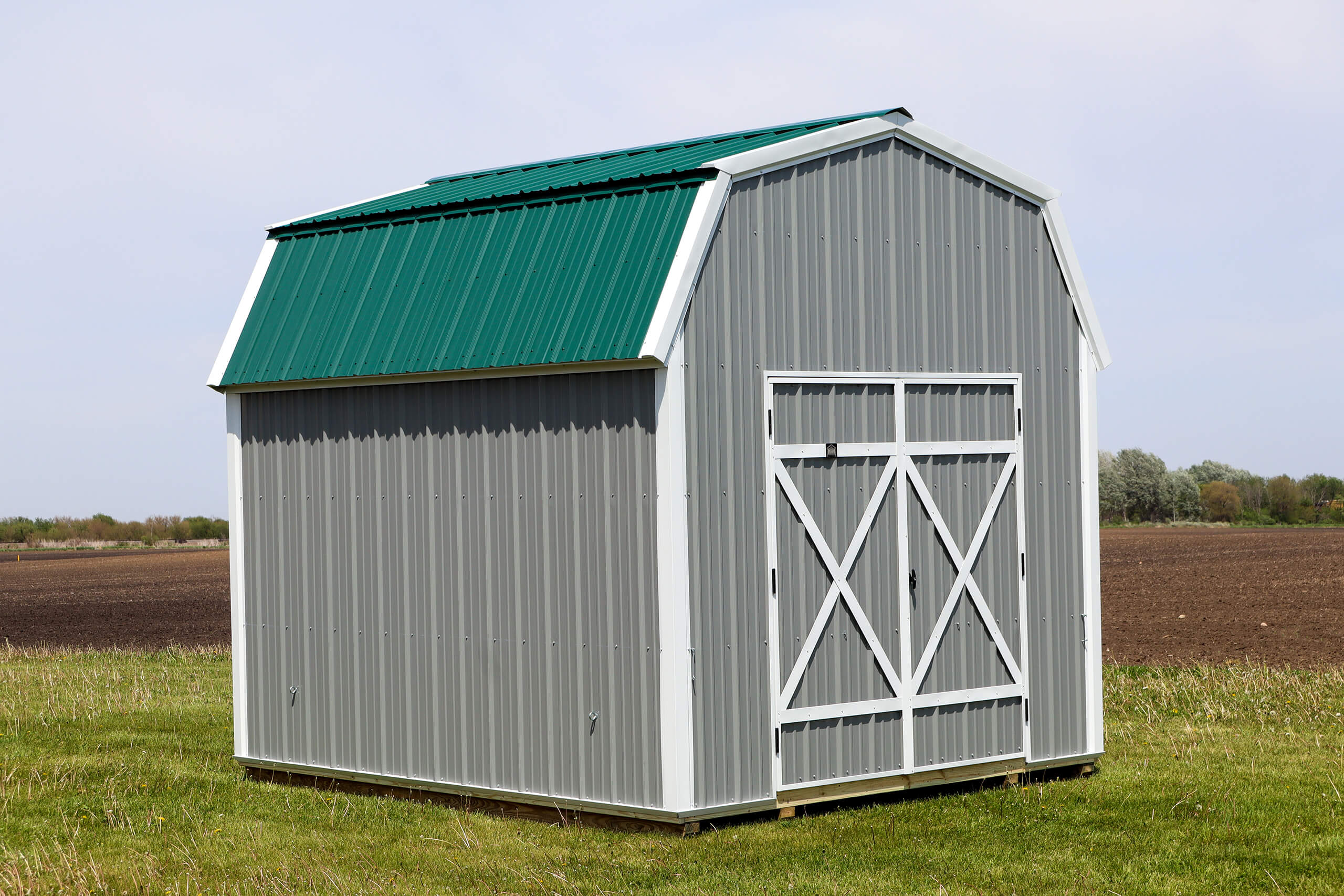 A small gray metal barn with a green roof stands on grass beside a plowed field under a partly cloudy sky.