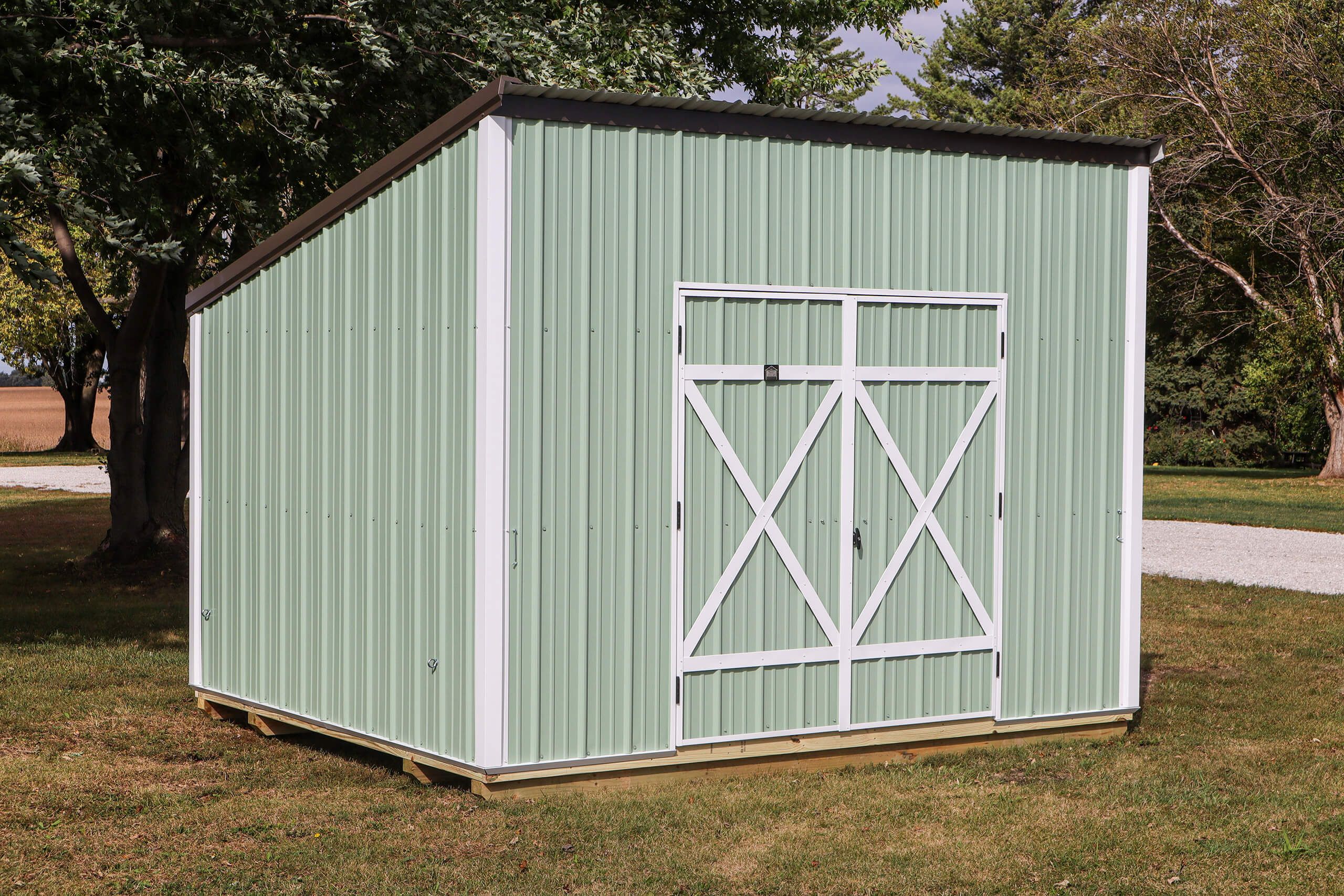 A light green metal storage shed with a sloped roof and double doors stands on a grassy lawn near trees.