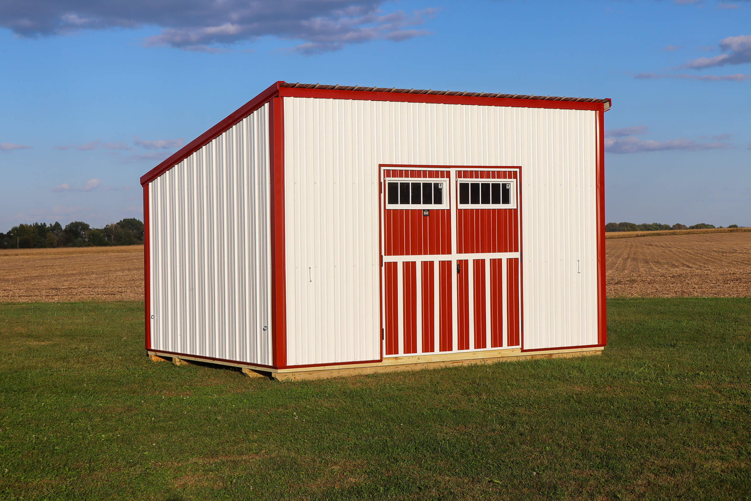 A red and white metal shed with double doors stands on a grassy field, with a plowed field and blue sky in the background.