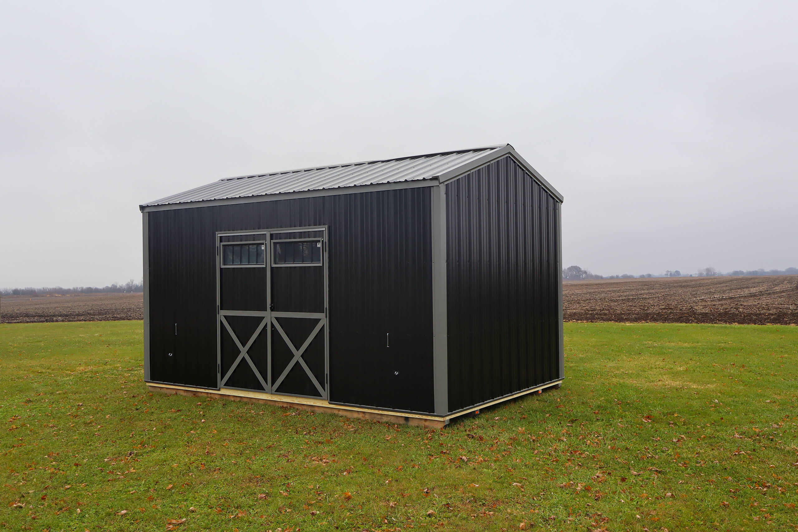 A small black metal shed with double doors and windows stands on a grassy field near a plowed area, under an overcast sky.