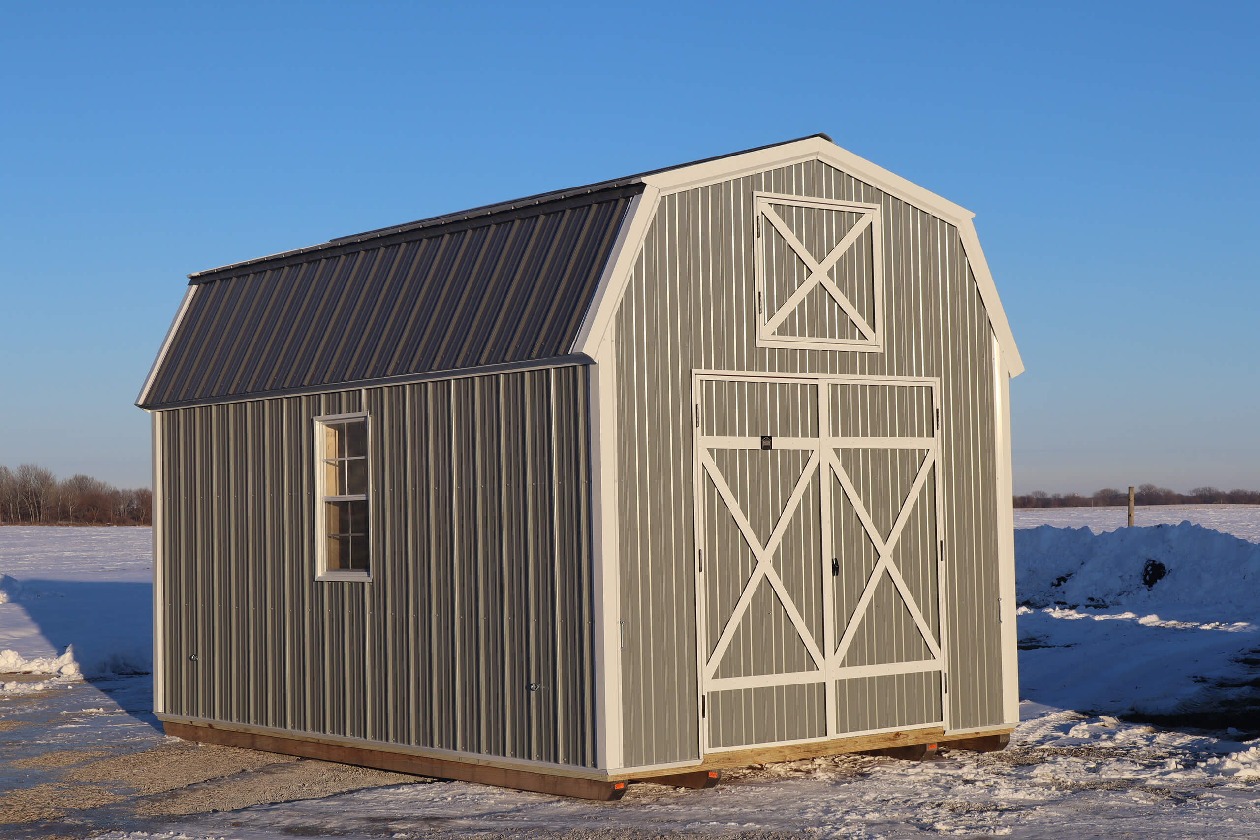 Gray metal barn-style shed with white trim sits on a snowy lot under a clear blue sky. The shed has double doors and a small window on the side.