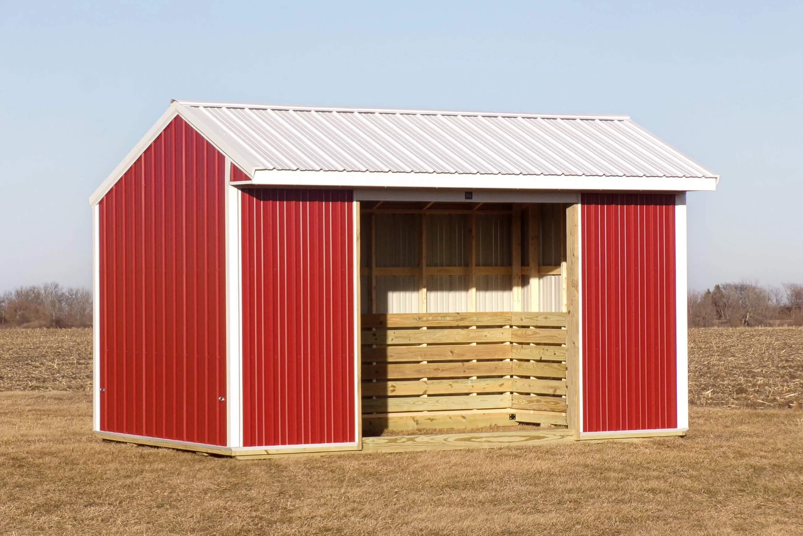 A small red and white shed with an open front sits on dry grass in a rural field under a clear sky.