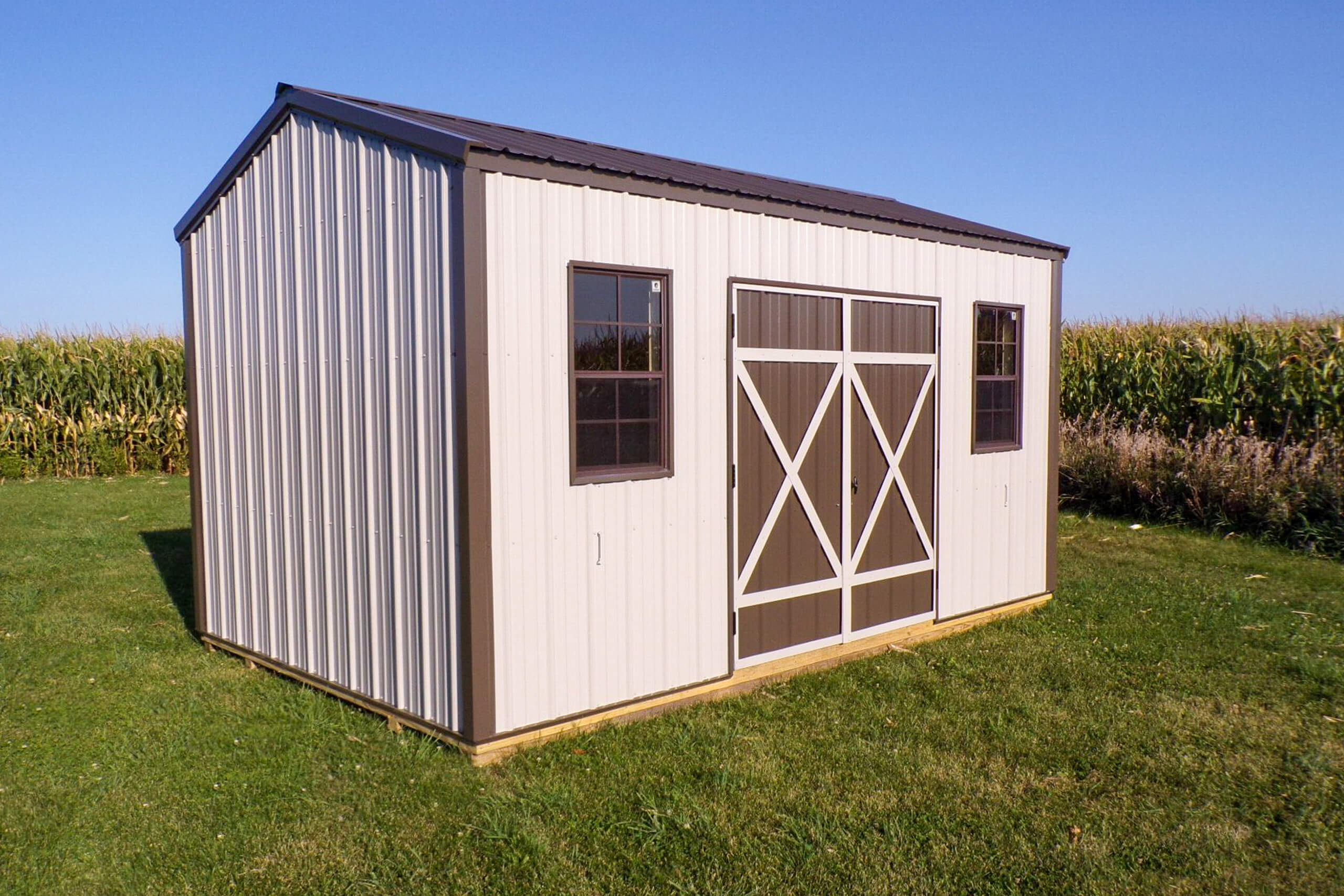 A metal storage shed with brown double doors and two windows stands on a grassy lawn, with a cornfield in the background under a clear blue sky.