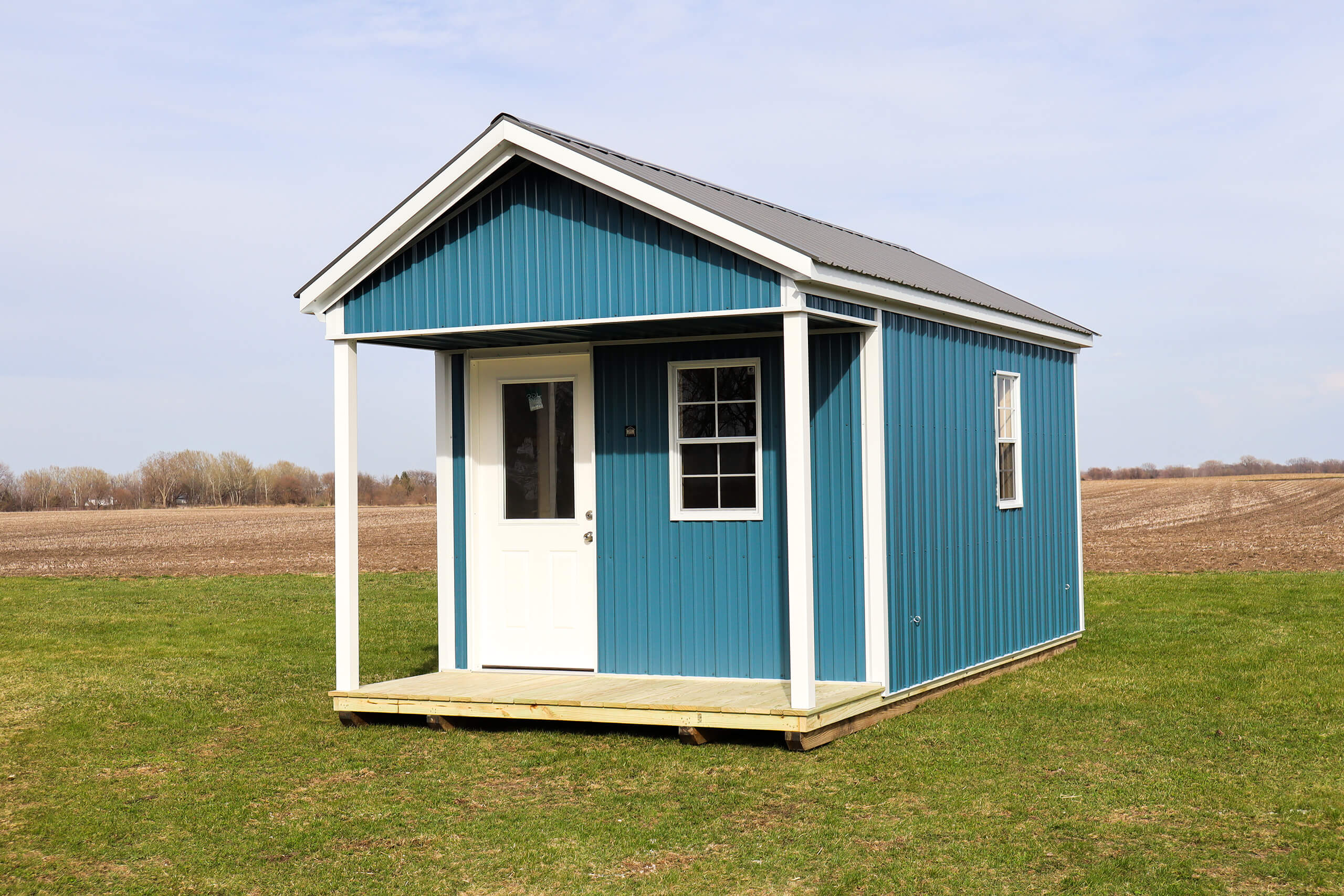 A small blue shed with white trim and a porch stands on a grassy field with bare farmland in the background under a partly cloudy sky.