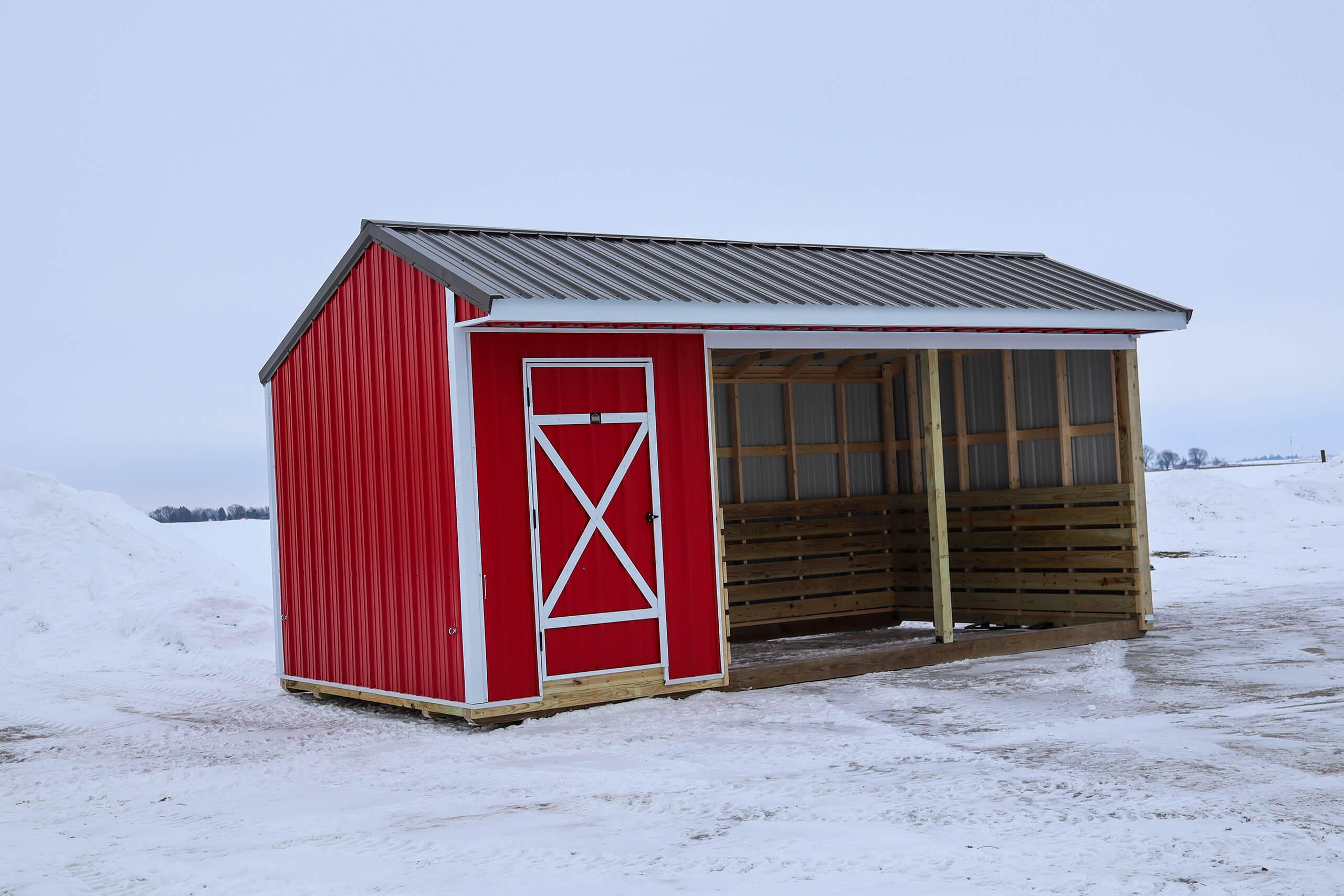 A red and white shed with a slanted metal roof stands on snowy ground. The shed has a closed section and an open, partially enclosed section.