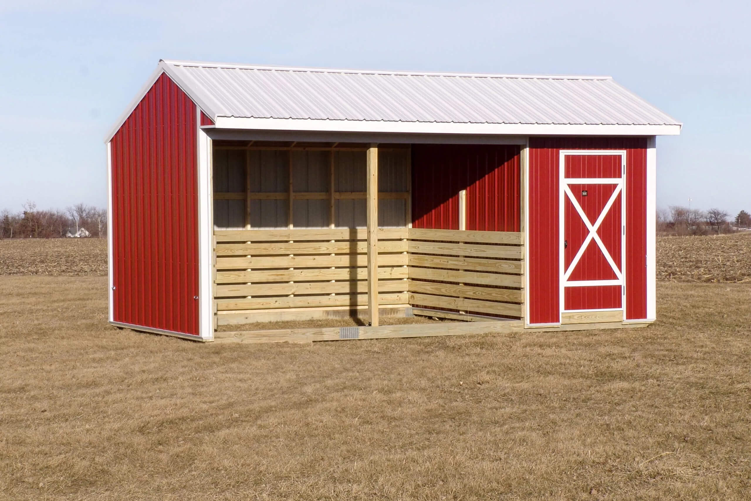 Red and white shed with a metal roof and open covered area, standing on dry grass in an empty field.