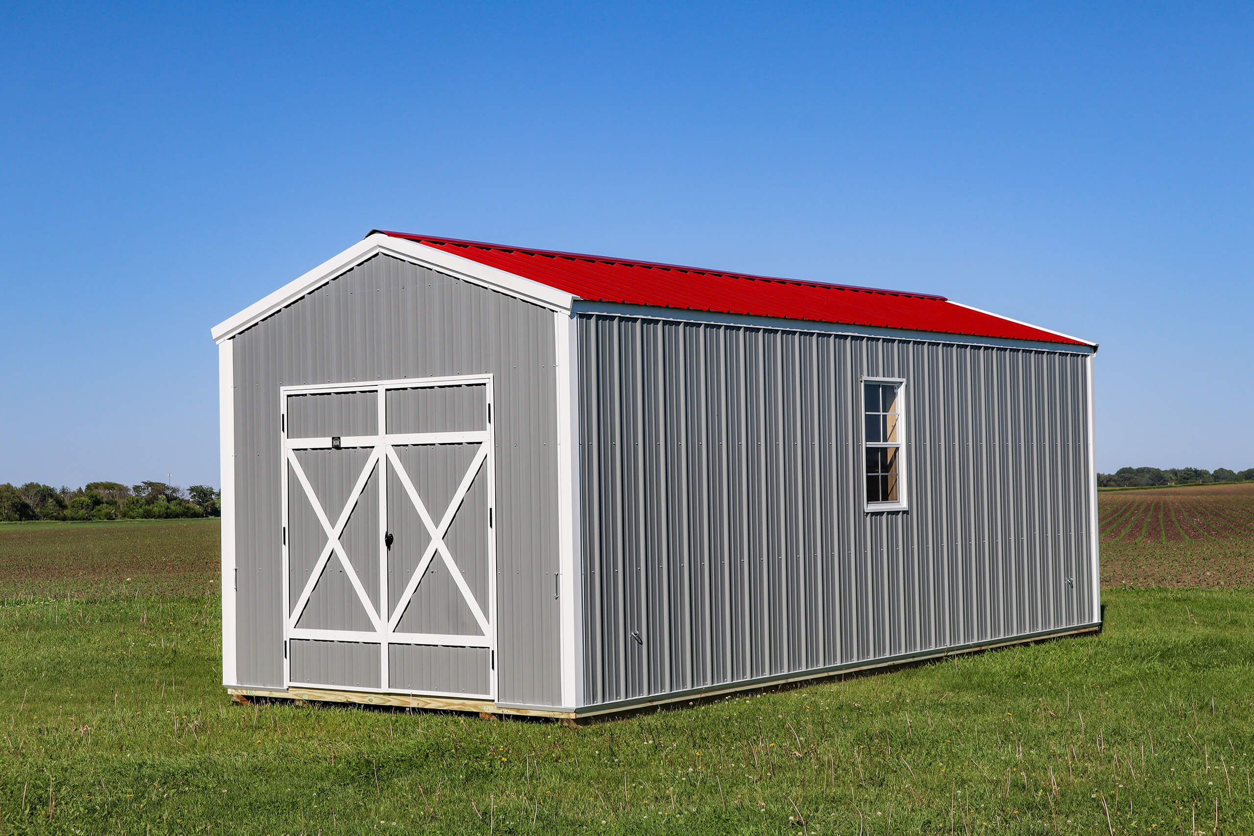 A gray metal shed with a red roof and white double doors stands on grass in an open field under a clear blue sky.