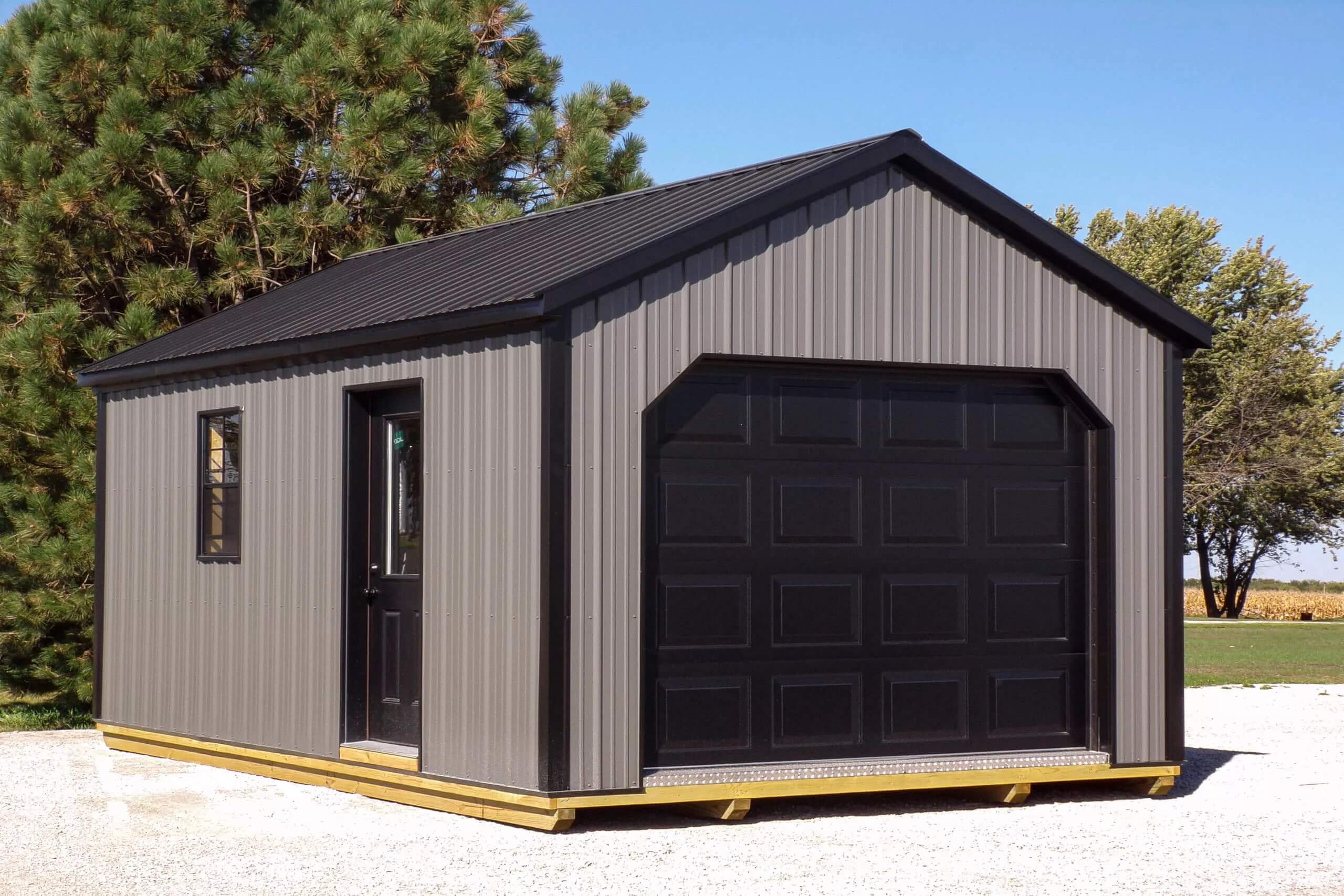 A small gray metal shed with a black garage door and side entry door sits on a gravel lot next to trees under a clear blue sky.