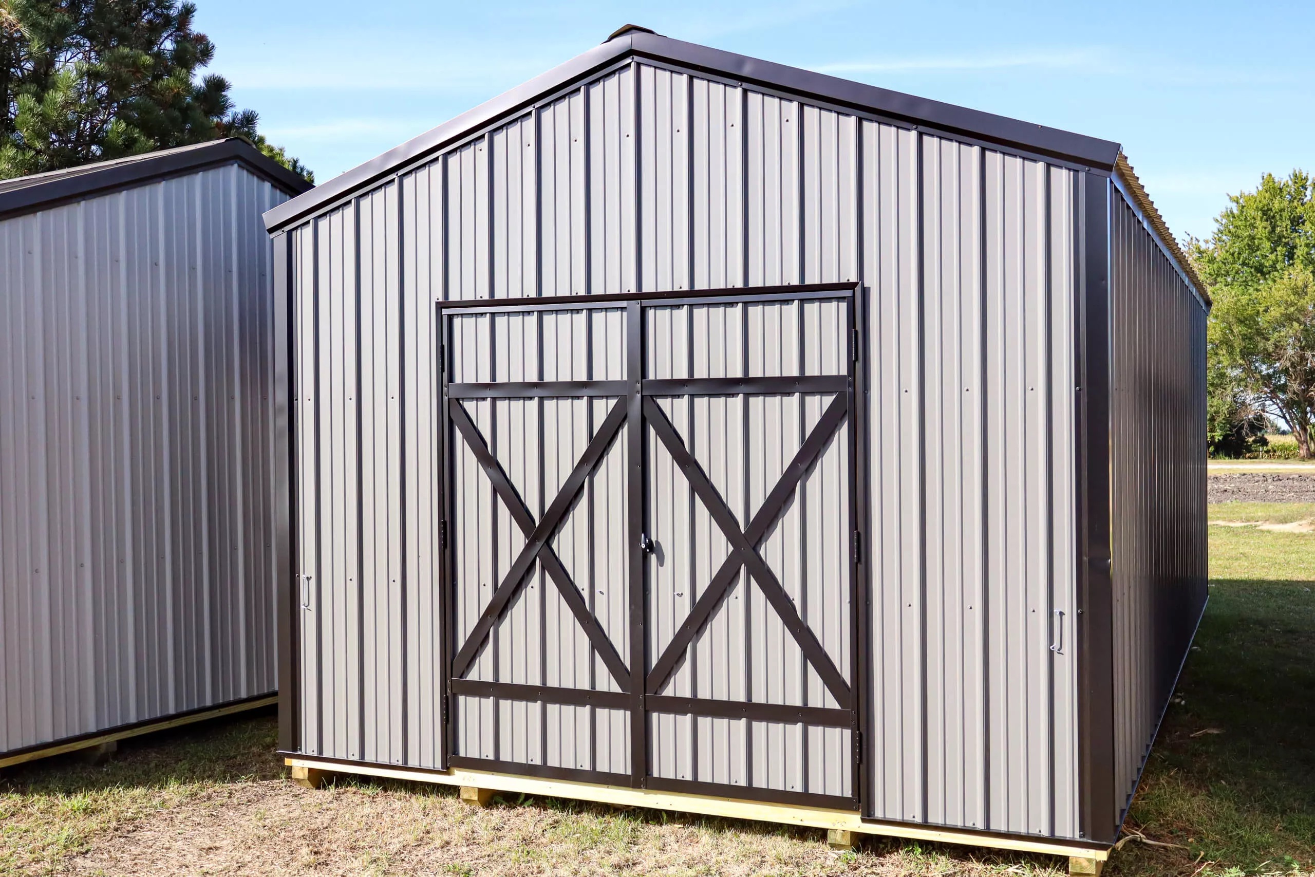 Gray metal storage shed with double doors, black trim, and a gable roof, standing on a grassy area with trees visible in the background.