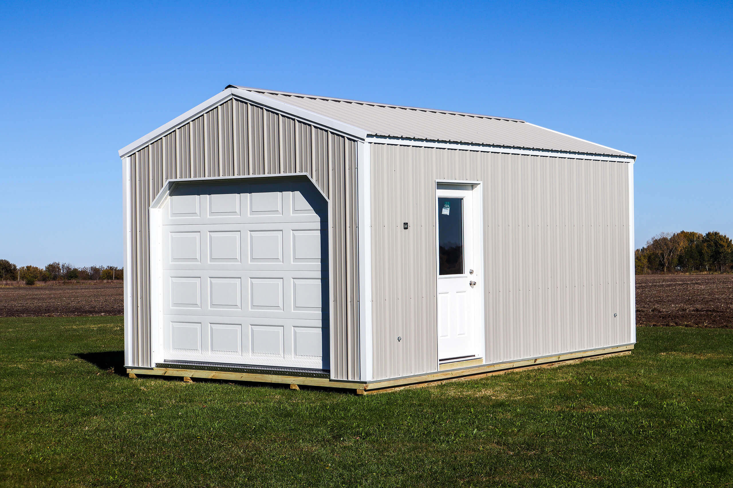 A small beige metal shed with a white garage door and a white entry door sits on a grassy field under a clear blue sky.