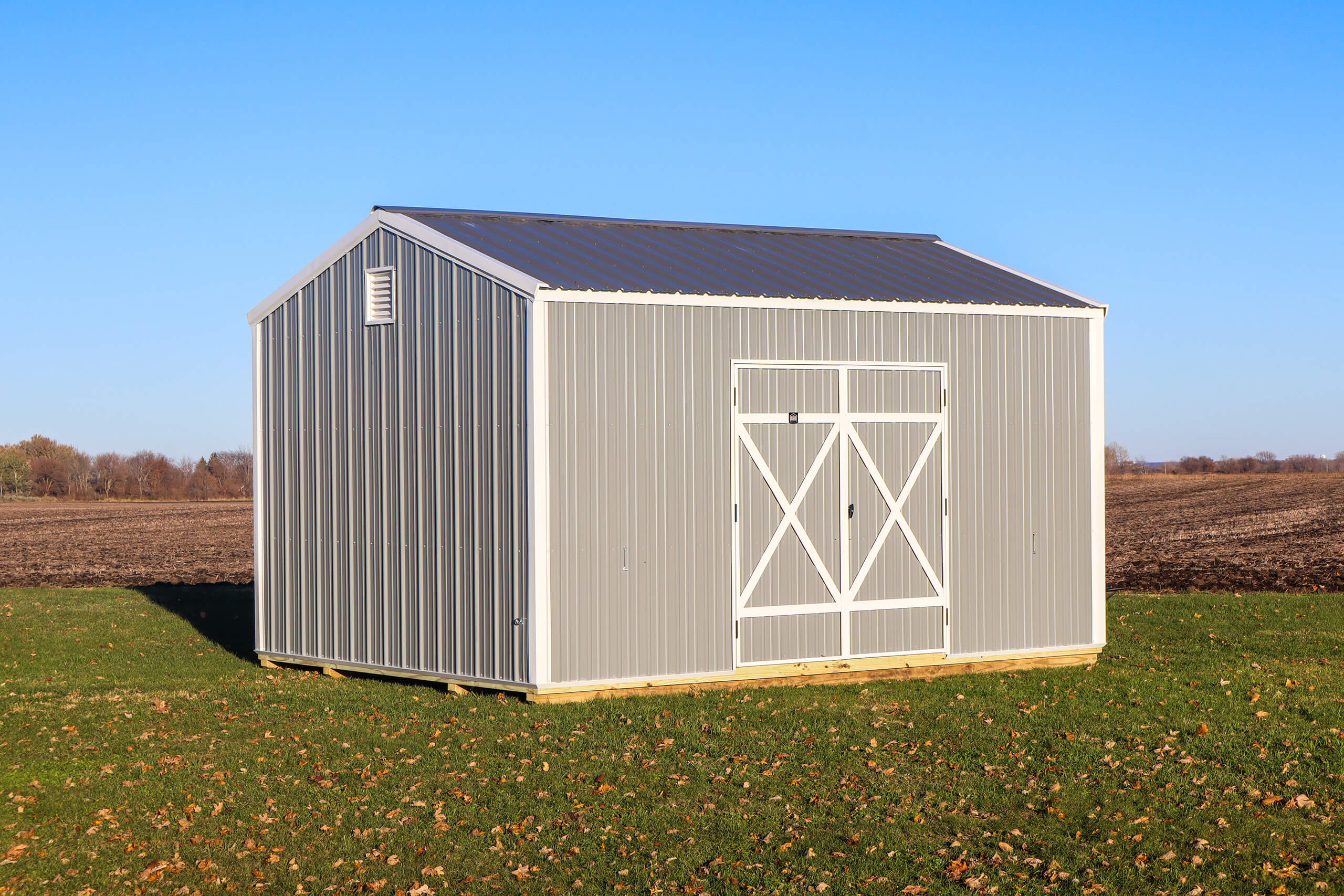 A gray metal storage shed with a white-trimmed double door stands on a grassy area beside a plowed field under a clear blue sky.