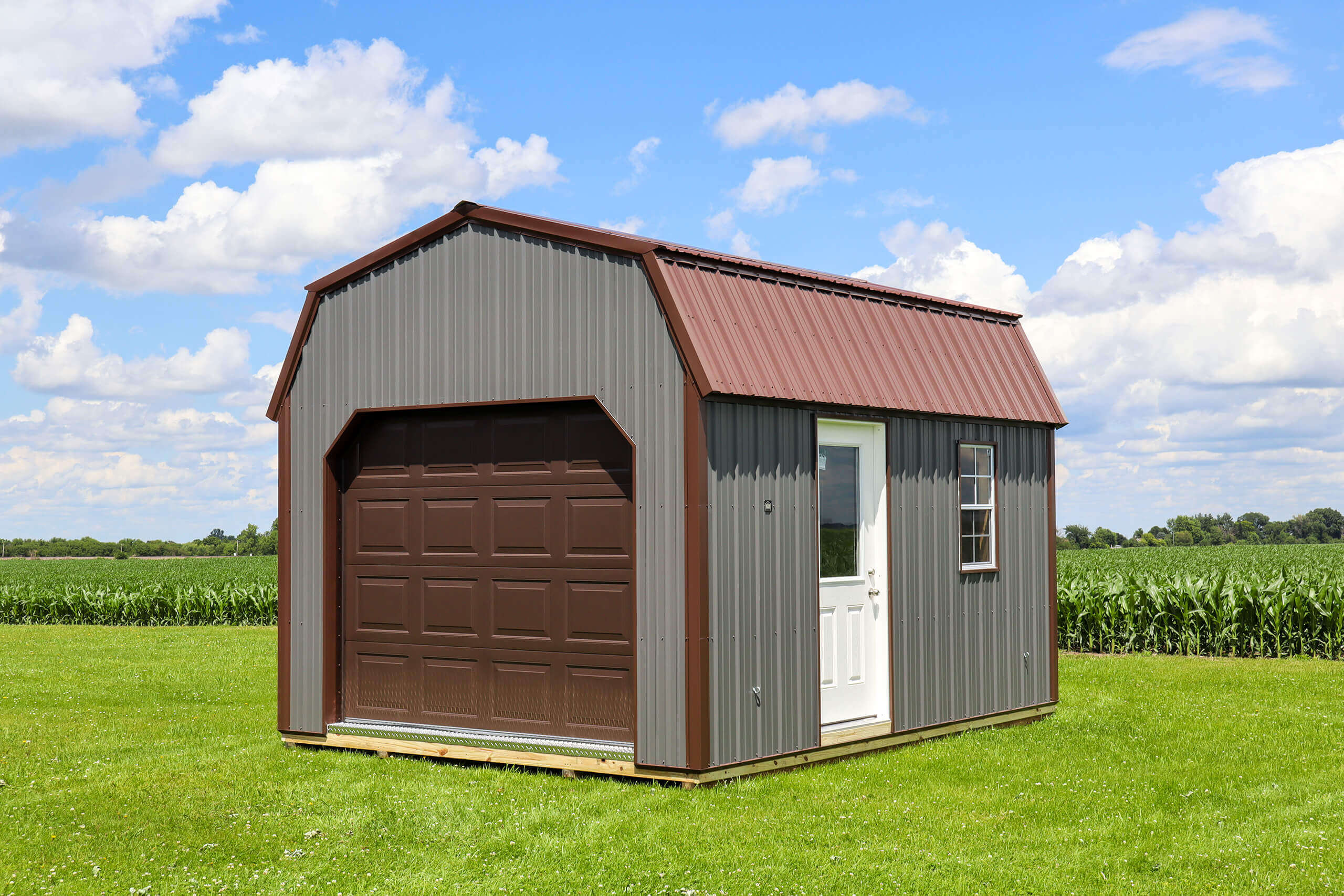 A small gray and brown metal storage shed with a garage door, white entry door, and window, sitting on green grass near a cornfield under a blue sky with clouds.