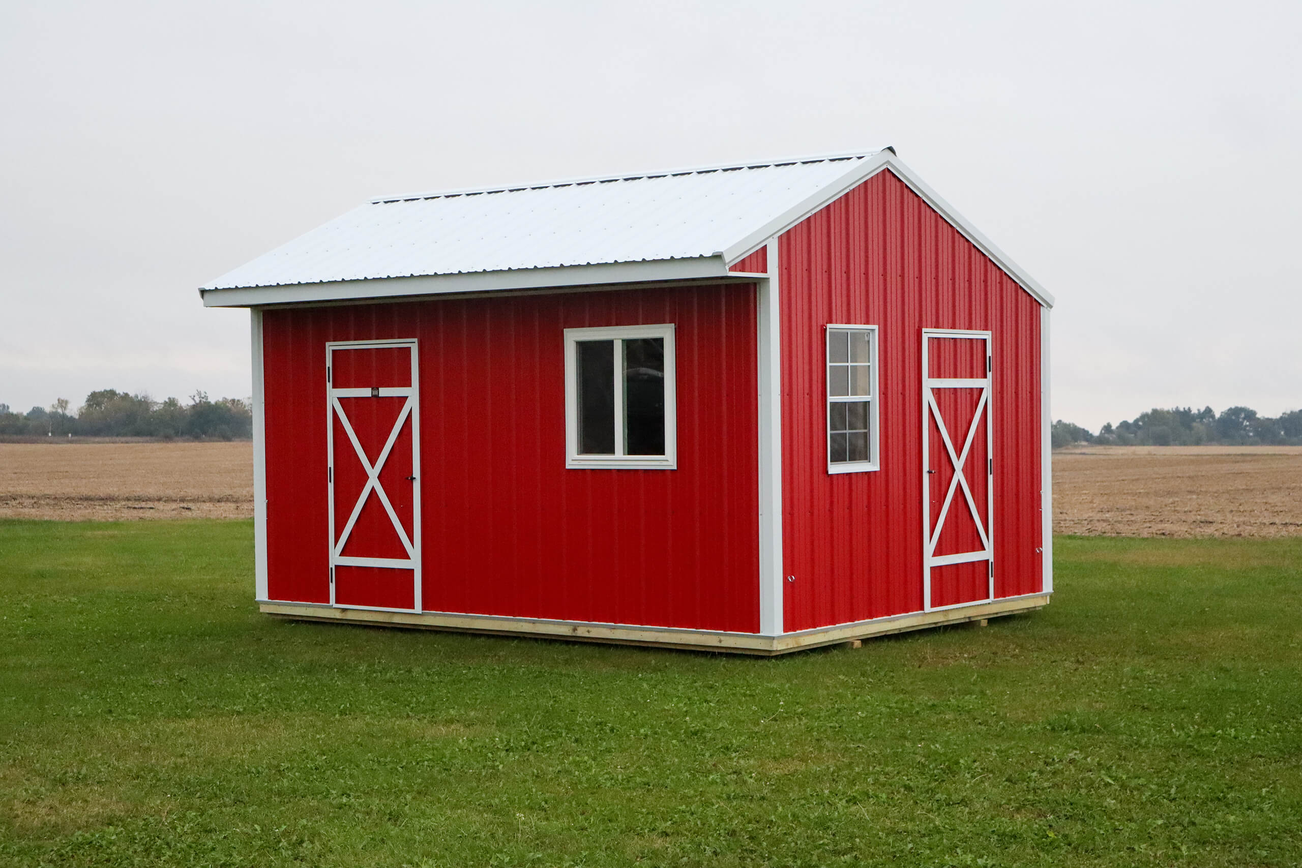 A small red shed with a white metal roof, two white-trimmed doors, and one window sits on a grassy field with farmland in the background.