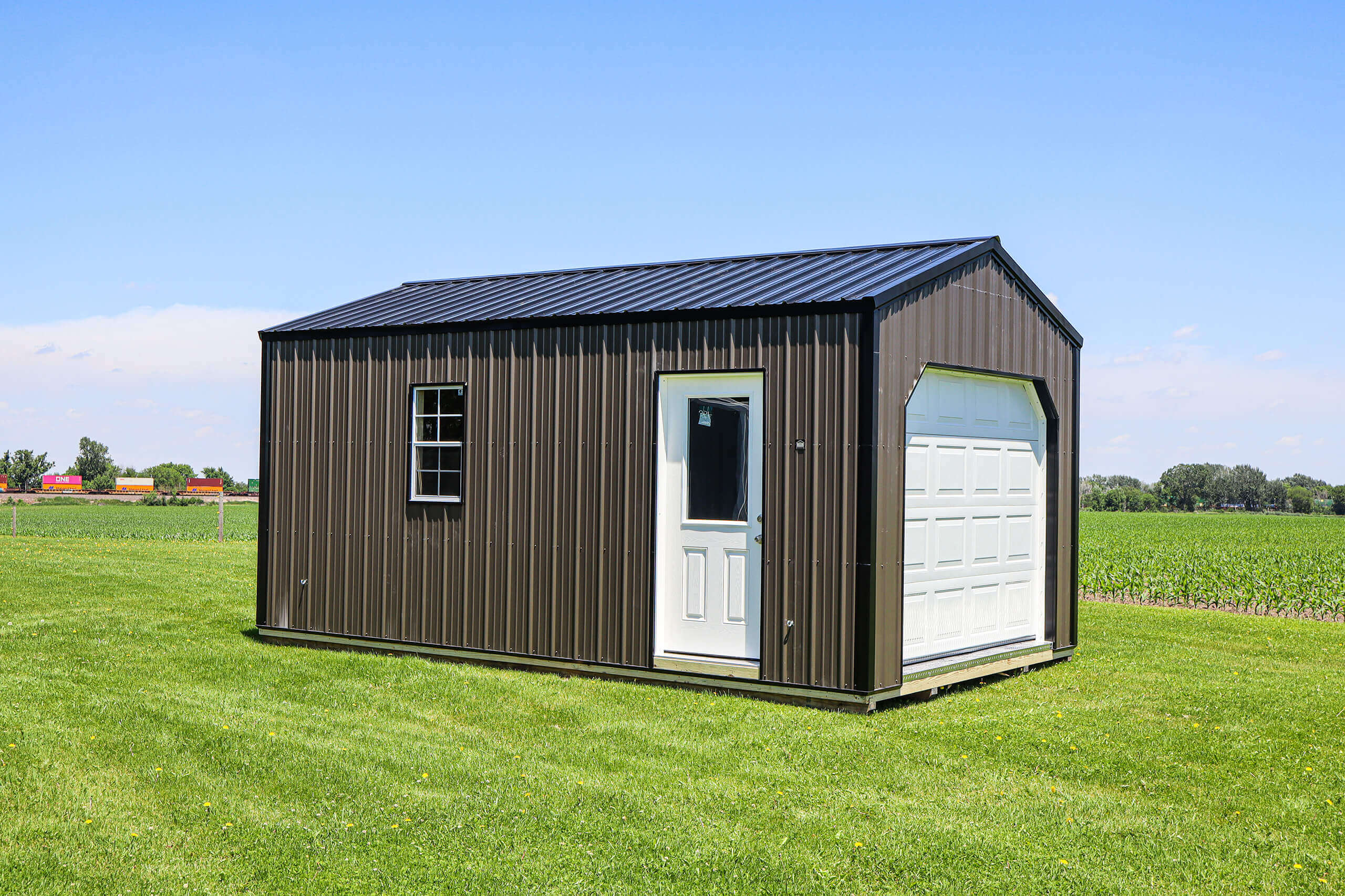 A small metal shed with a window, white door, and garage door sits on a grassy lawn near a field under a clear blue sky.