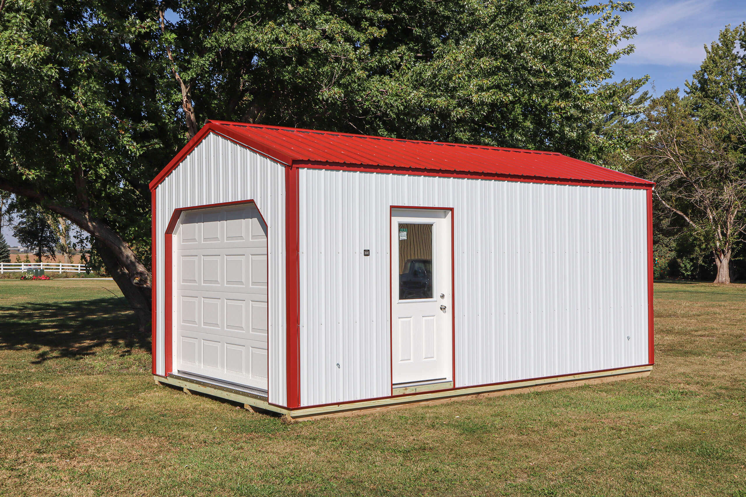 Small white metal storage shed with a red roof, one garage door, and one entry door, situated on a grassy lawn with trees in the background.