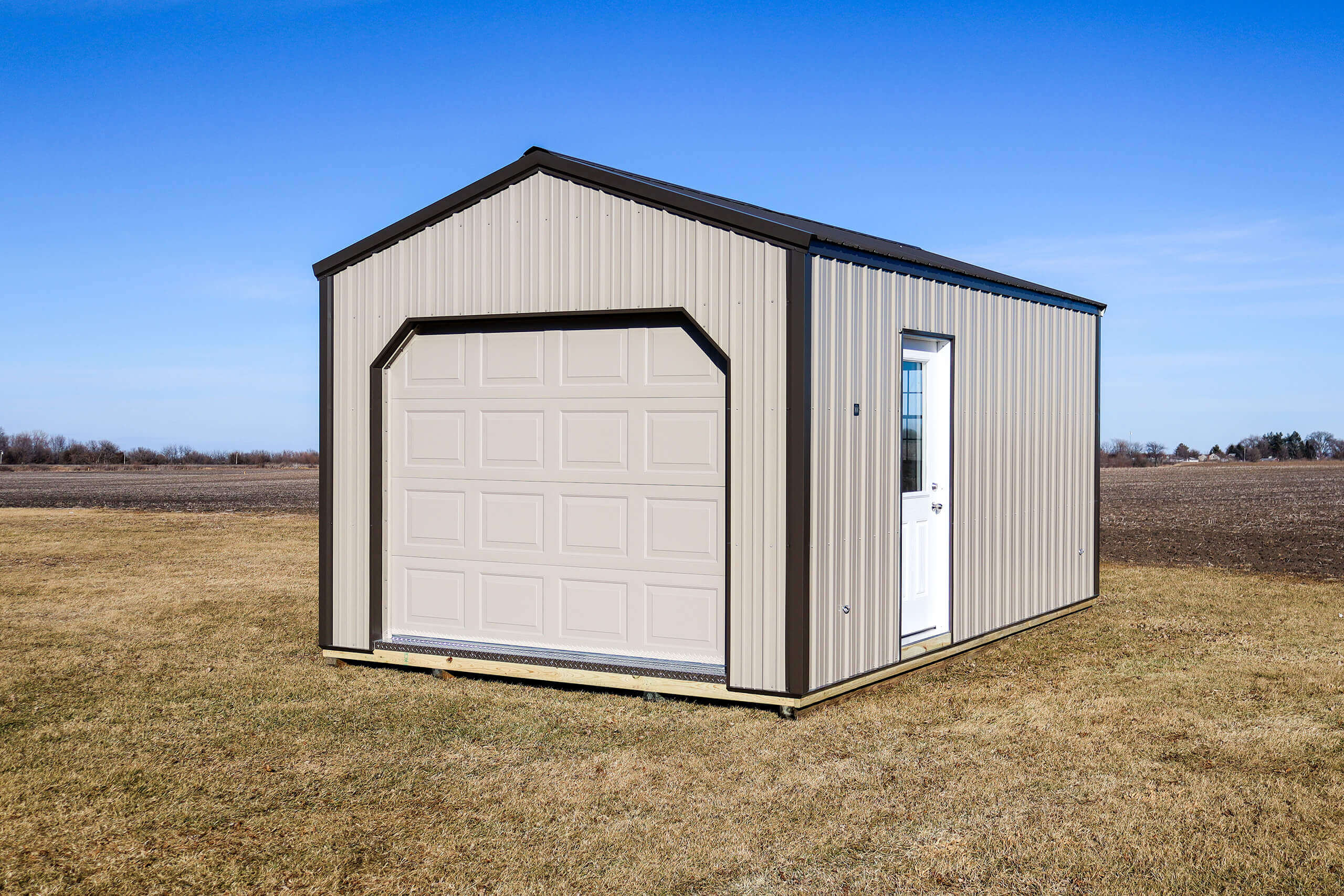 A small beige metal shed with a garage door and a white side door sits on a grassy field under a clear blue sky.