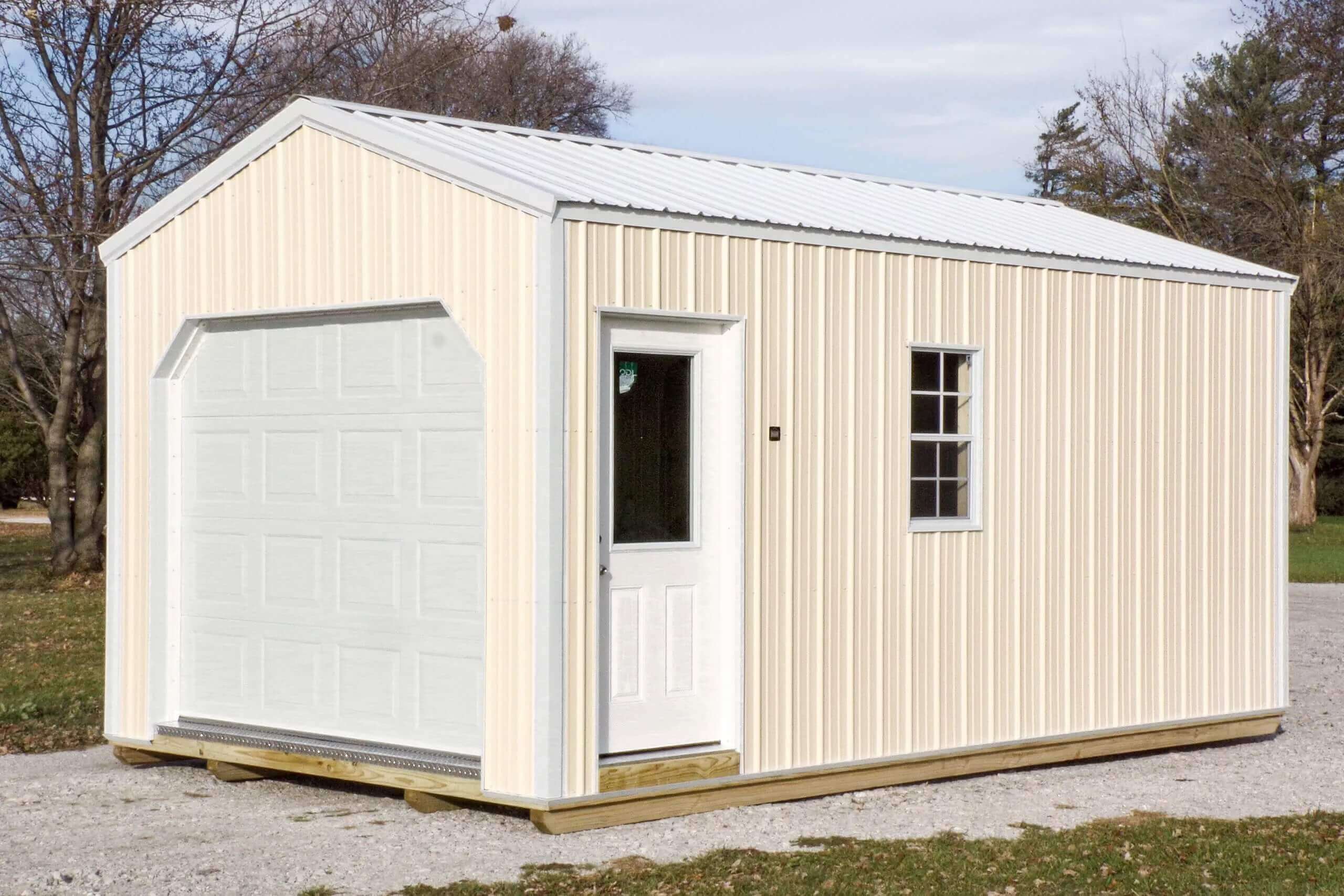 Small cream-colored metal shed with a white garage door, single entry door with window, and one side window, situated outdoors on a gravel area, surrounded by grass and trees.
