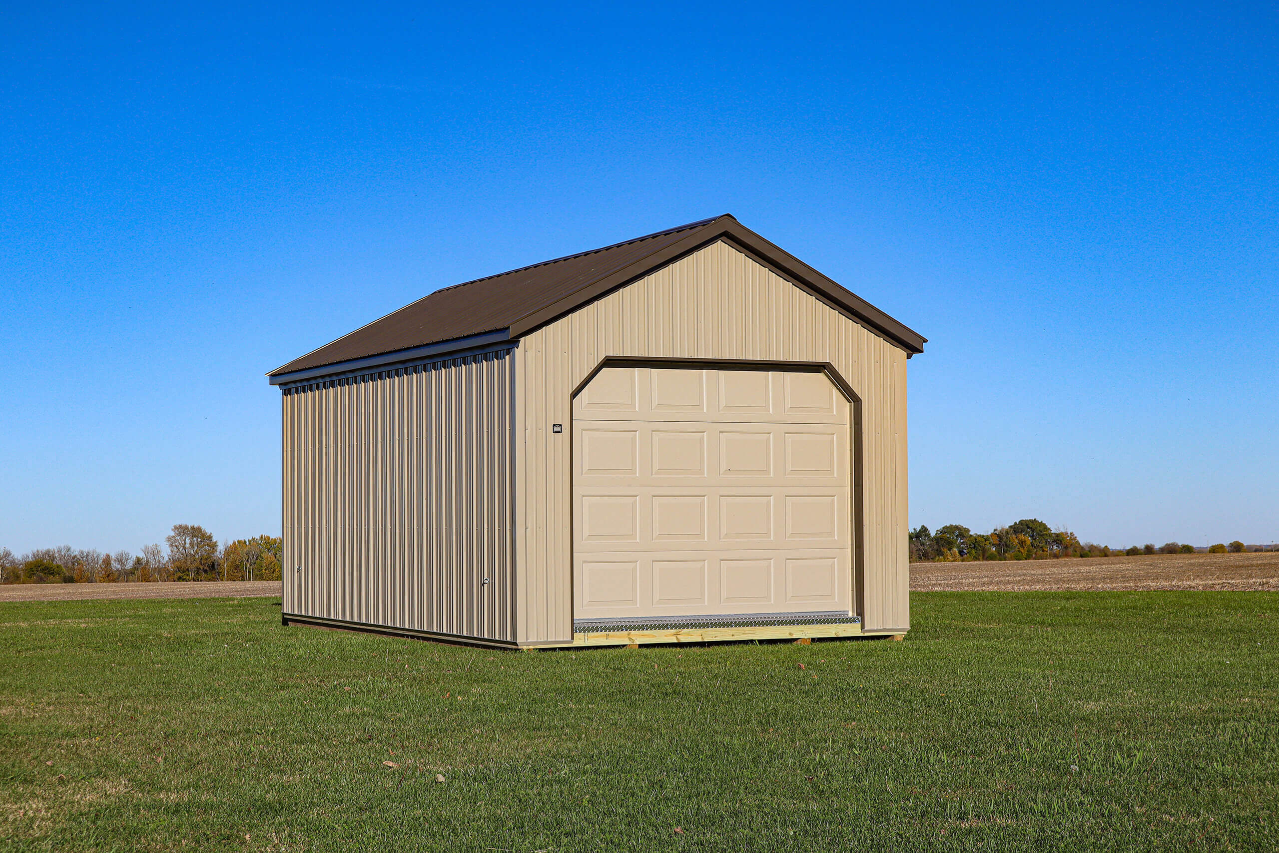 A beige metal shed with a closed garage door sits on green grass under a clear blue sky, with a field and trees in the background.