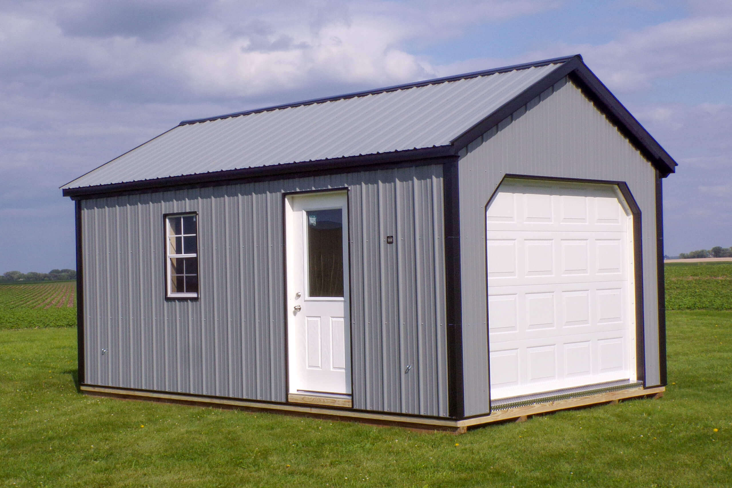 A small, gray metal shed with a white door, window, and garage door sits on a grassy field under a cloudy sky.