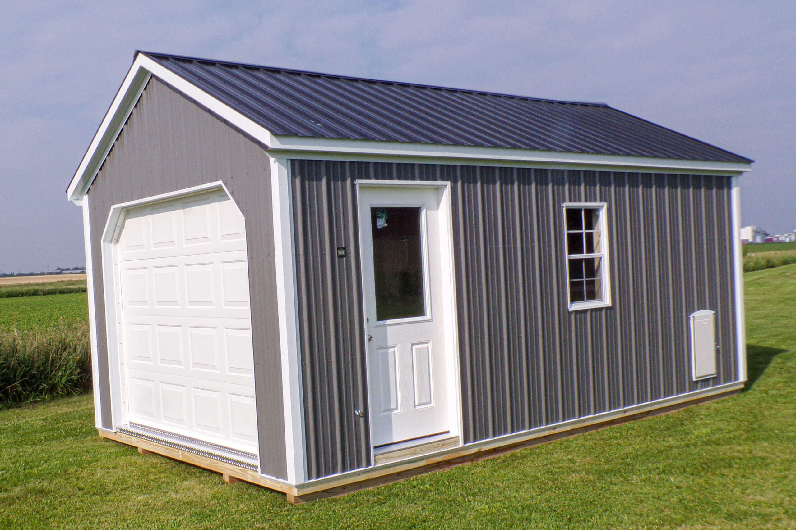 A small gray metal shed with a white garage door and entry door stands on a grassy field under a partly cloudy sky.