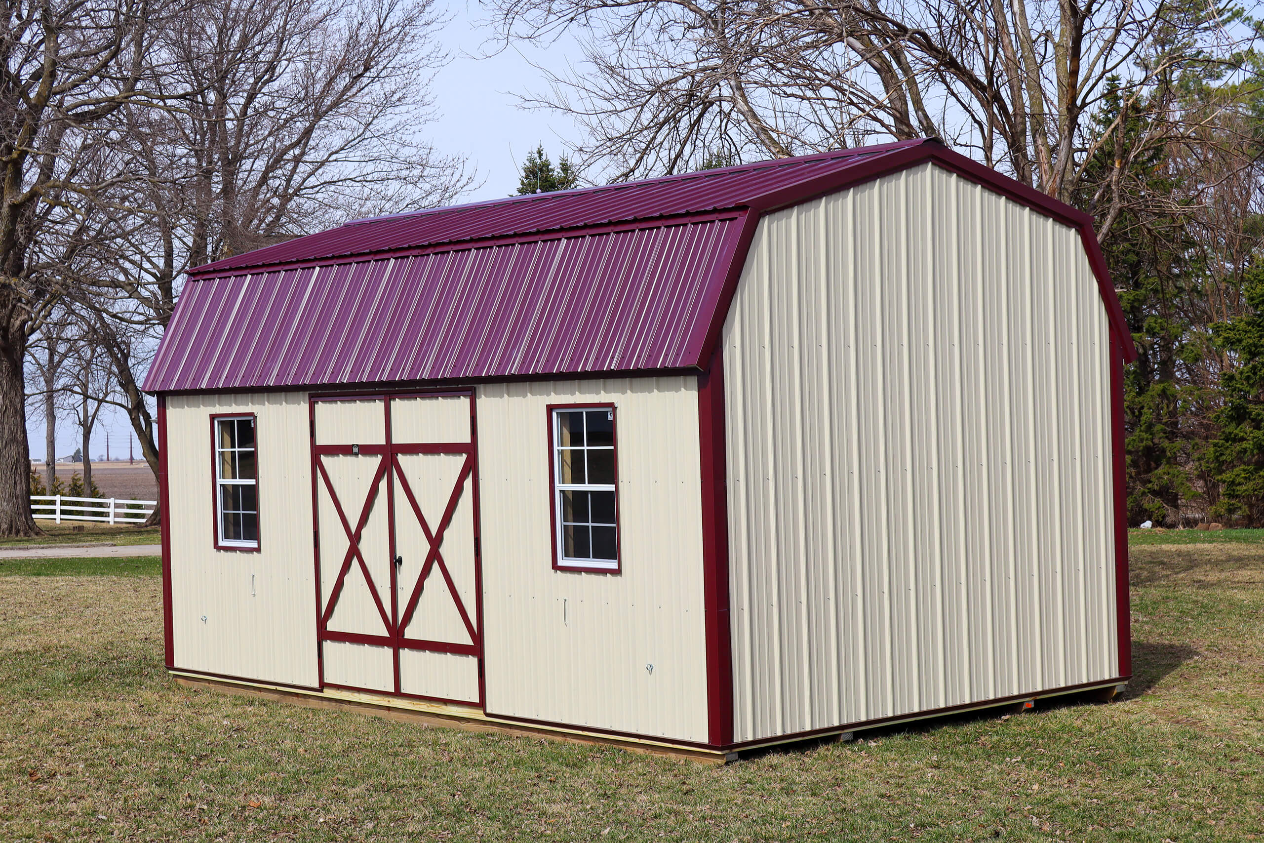 A beige metal storage shed with a red roof and trim sits on grass near leafless trees and a white fence.