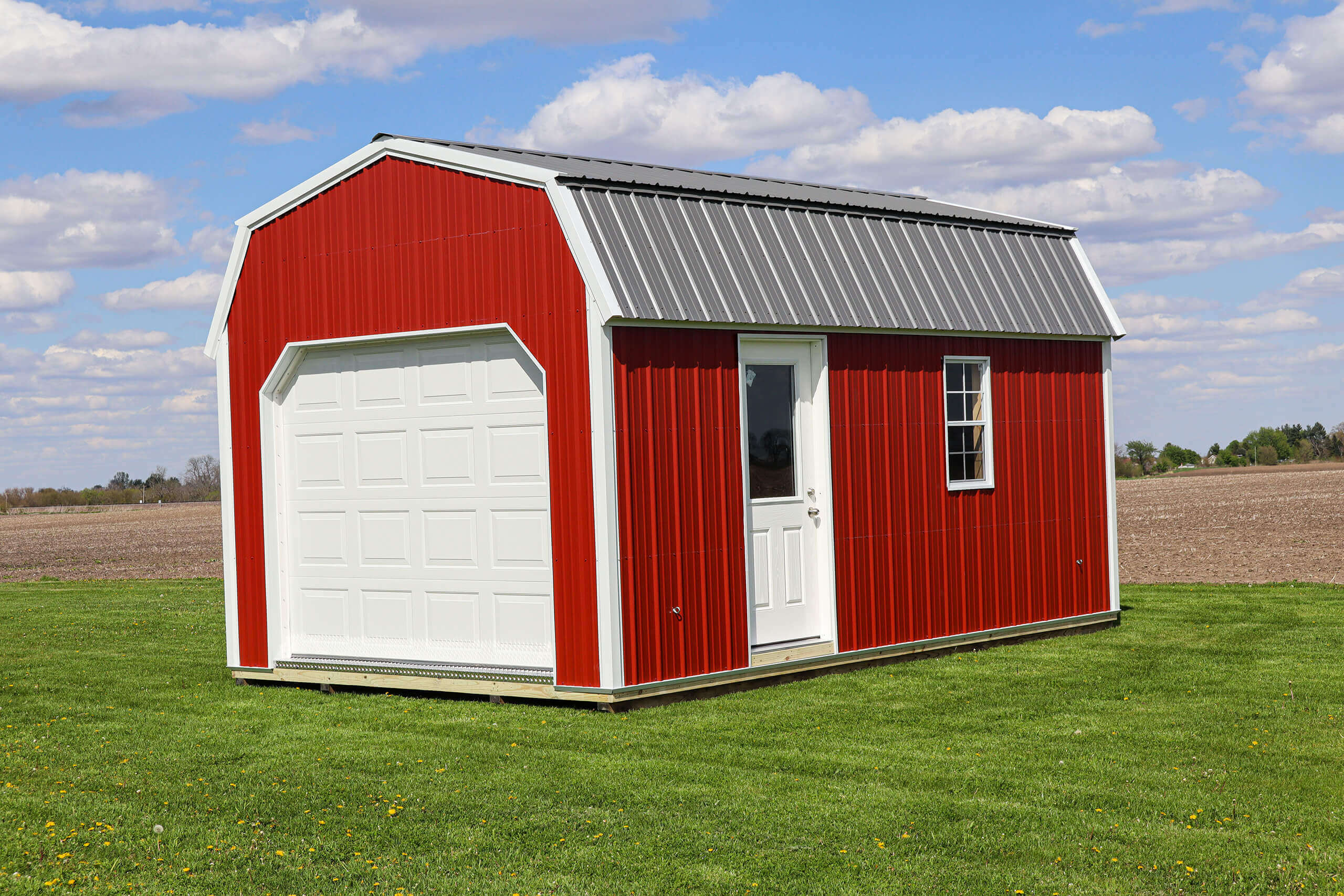 A small red shed with white trim, a garage door, a regular door, and one window sits on a grassy lawn with a field in the background.