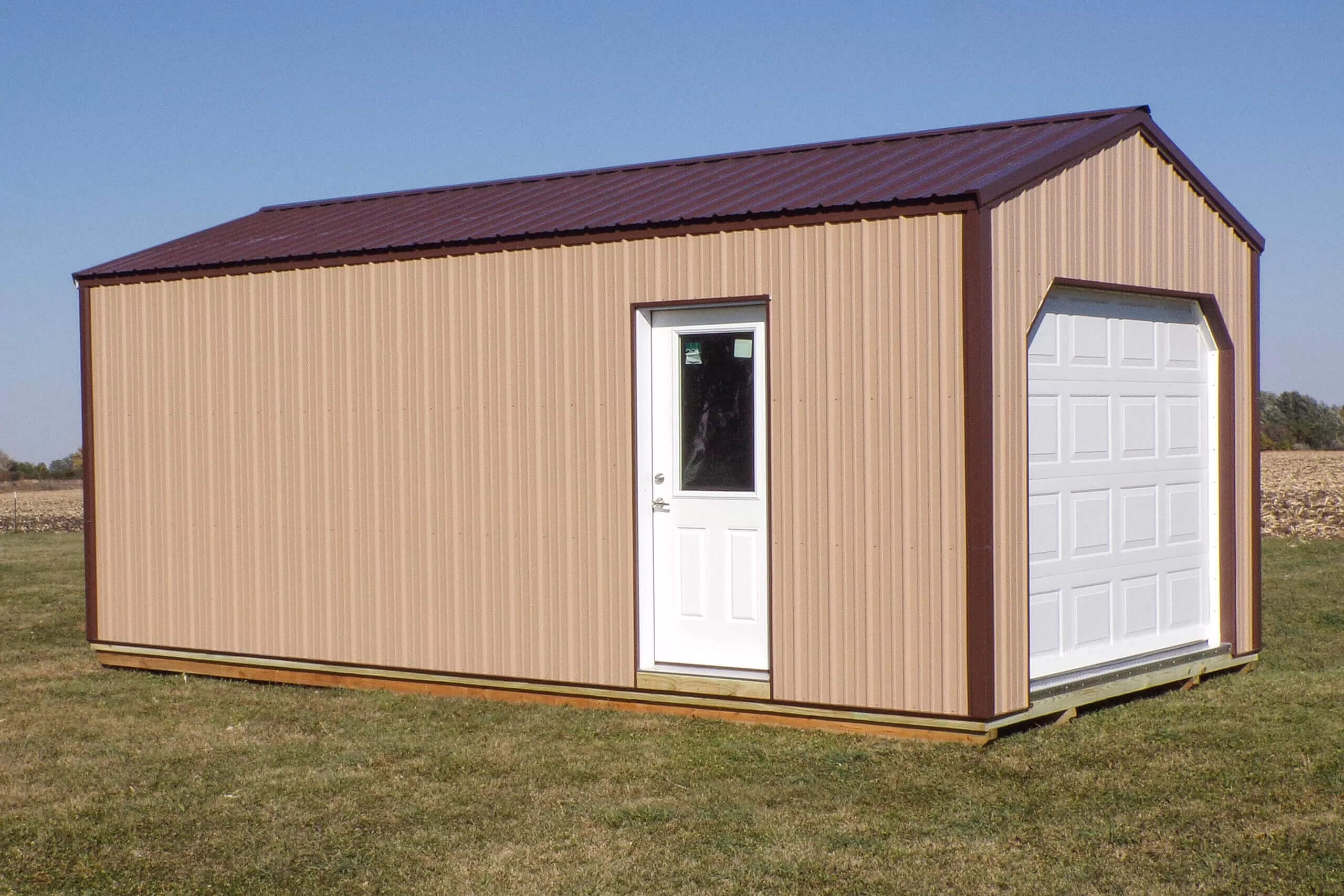 Small tan metal shed with a white side door and a white overhead garage door, sitting on a grassy field under a clear blue sky.