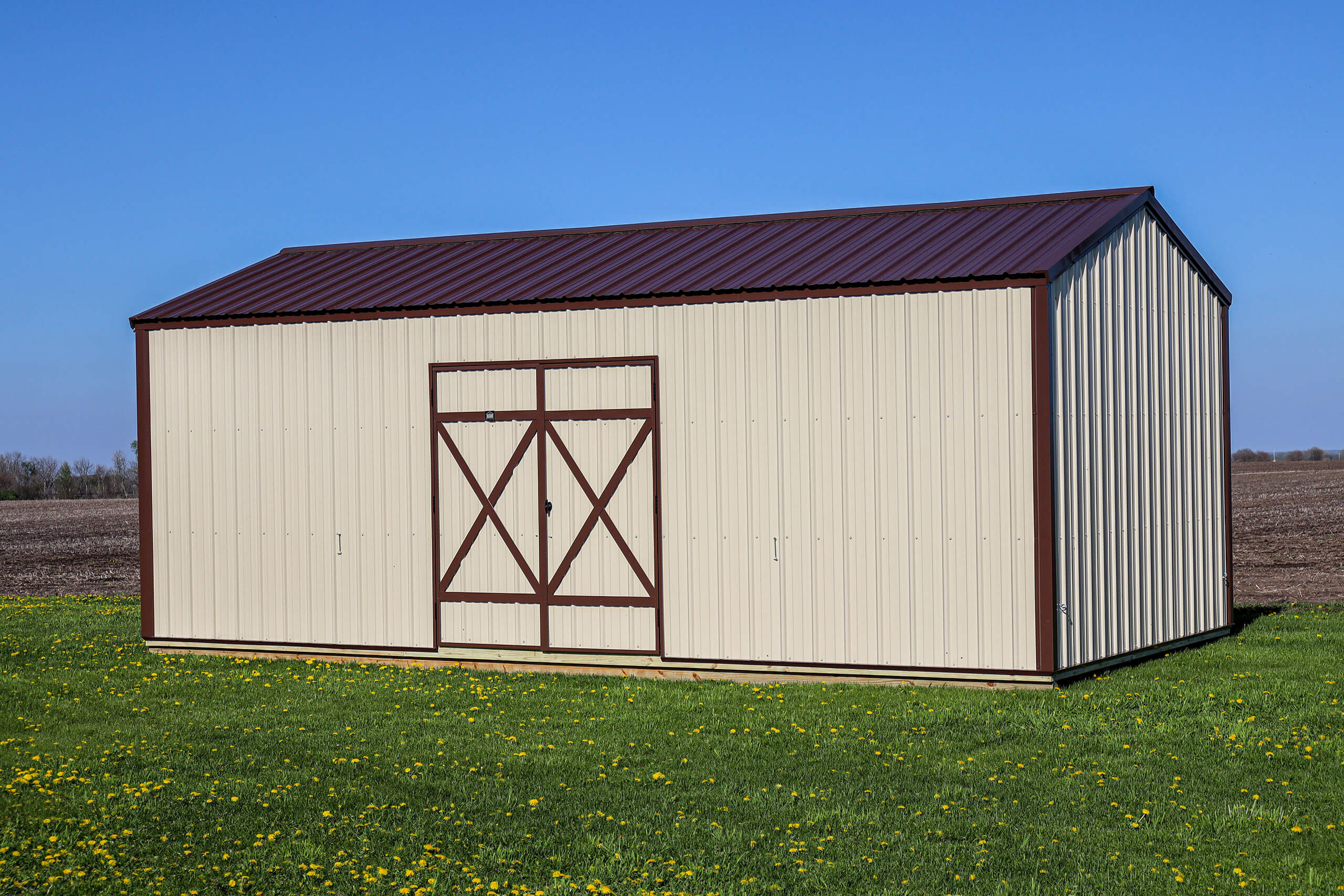A beige metal storage shed with brown trim and double doors stands on a grassy field under a clear blue sky.