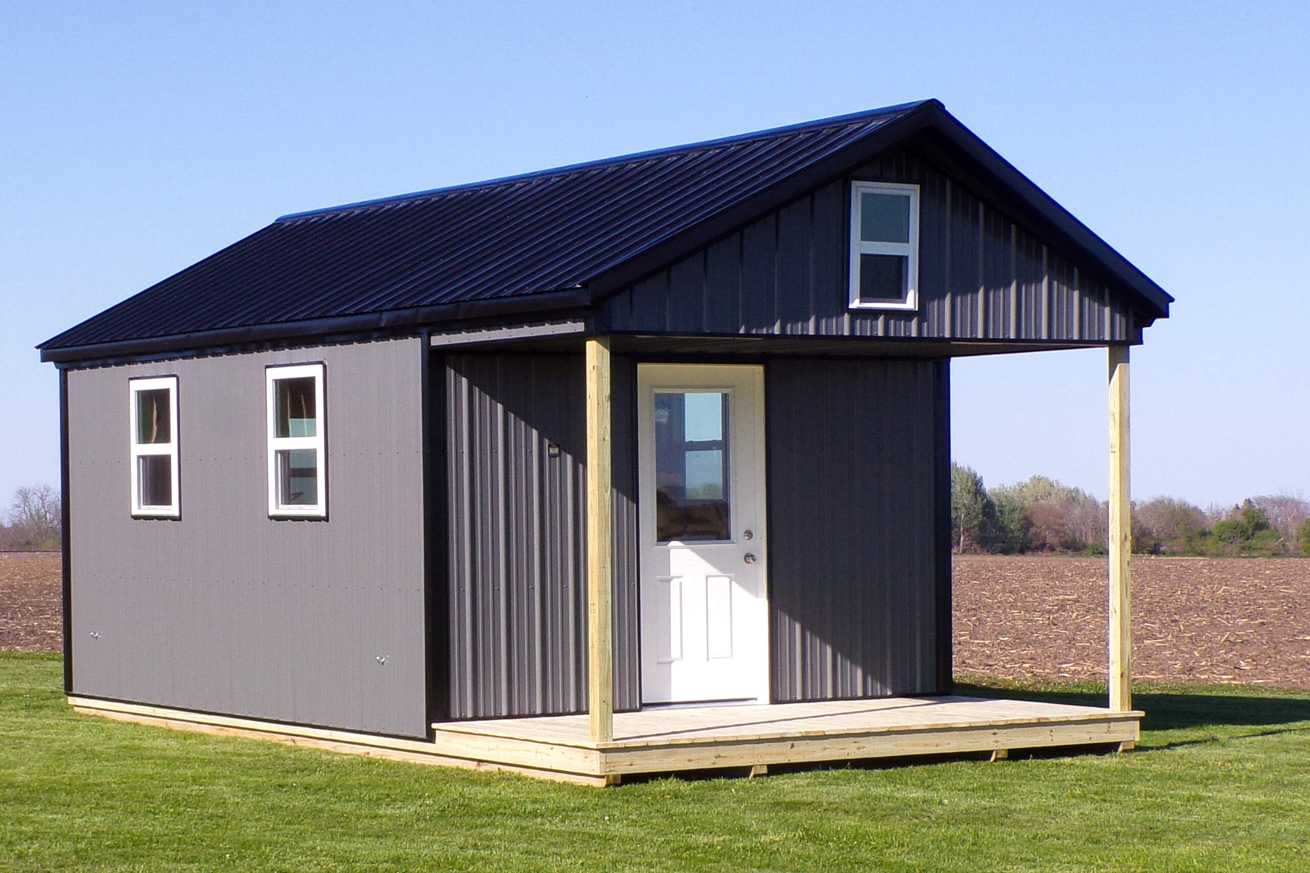 Small gray metal tiny house with a black roof, white door, two windows, and a small covered porch, situated on a grassy lawn with open field in the background.