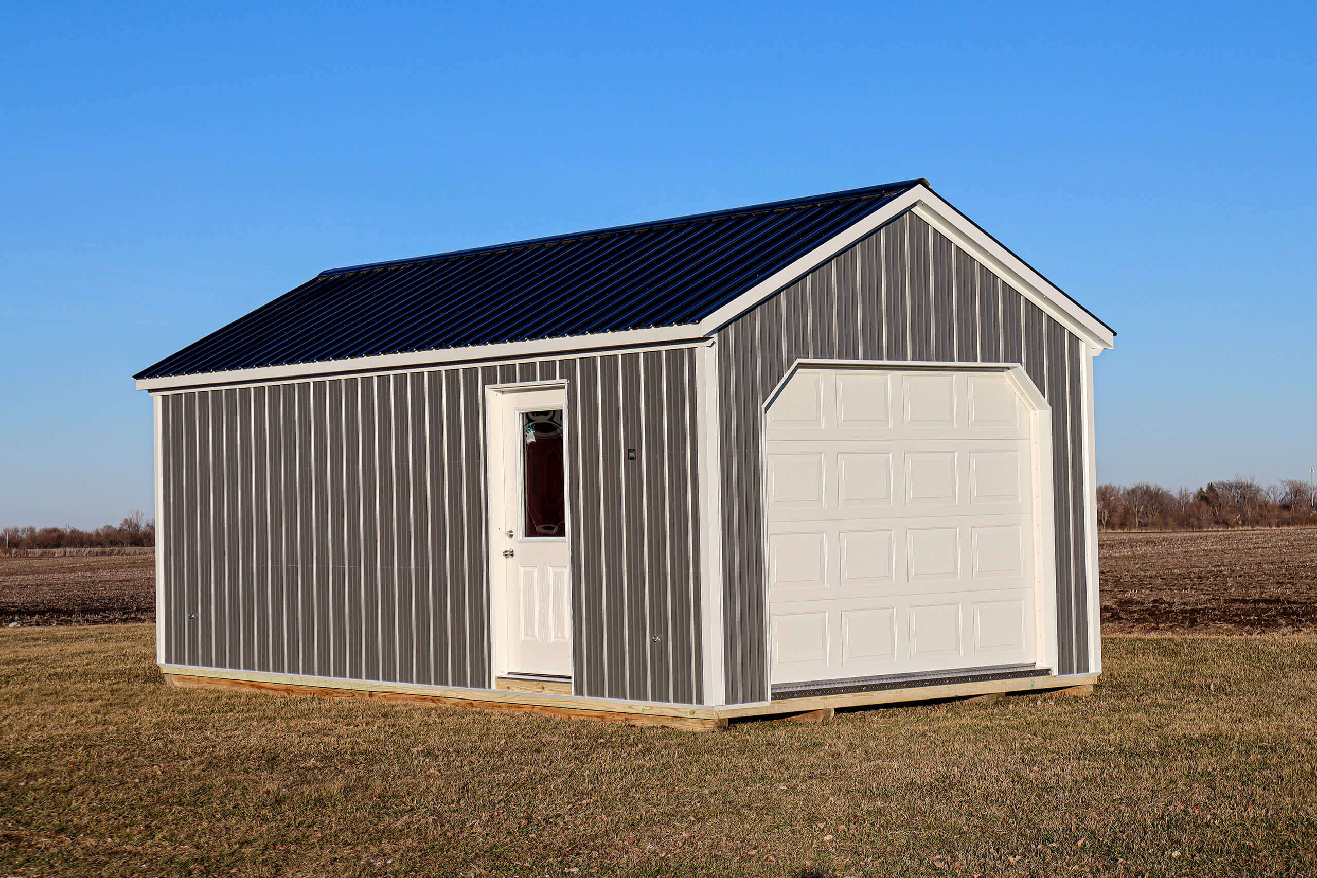 A small gray metal shed with a white door and a white garage door sits on grass in a field under a clear blue sky.