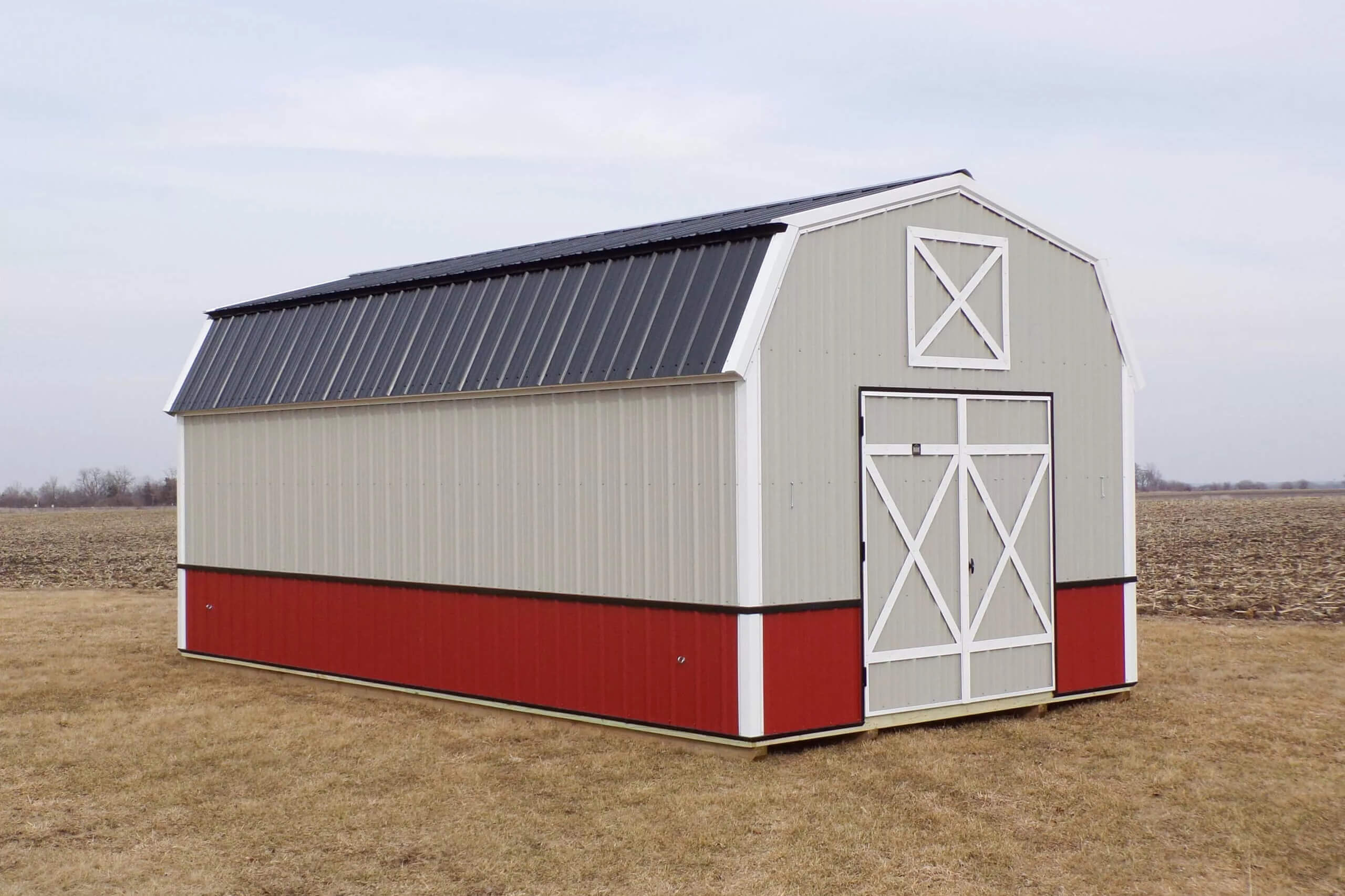 A red and beige barn-style shed with a black metal roof stands on a grassy field under a cloudy sky.