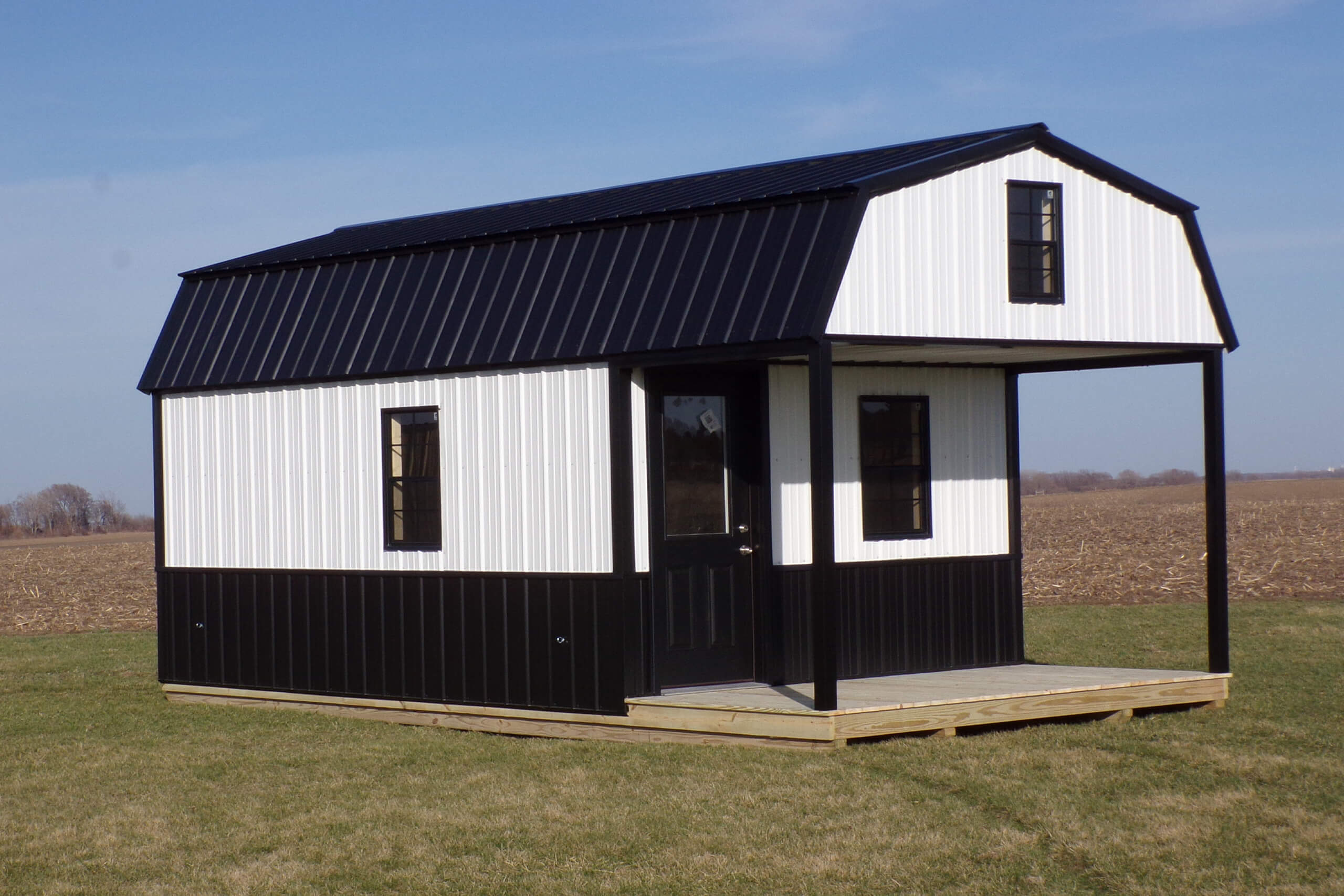 A small black and white barn-style building with a porch sits on a grassy area, with farmland and blue sky in the background.