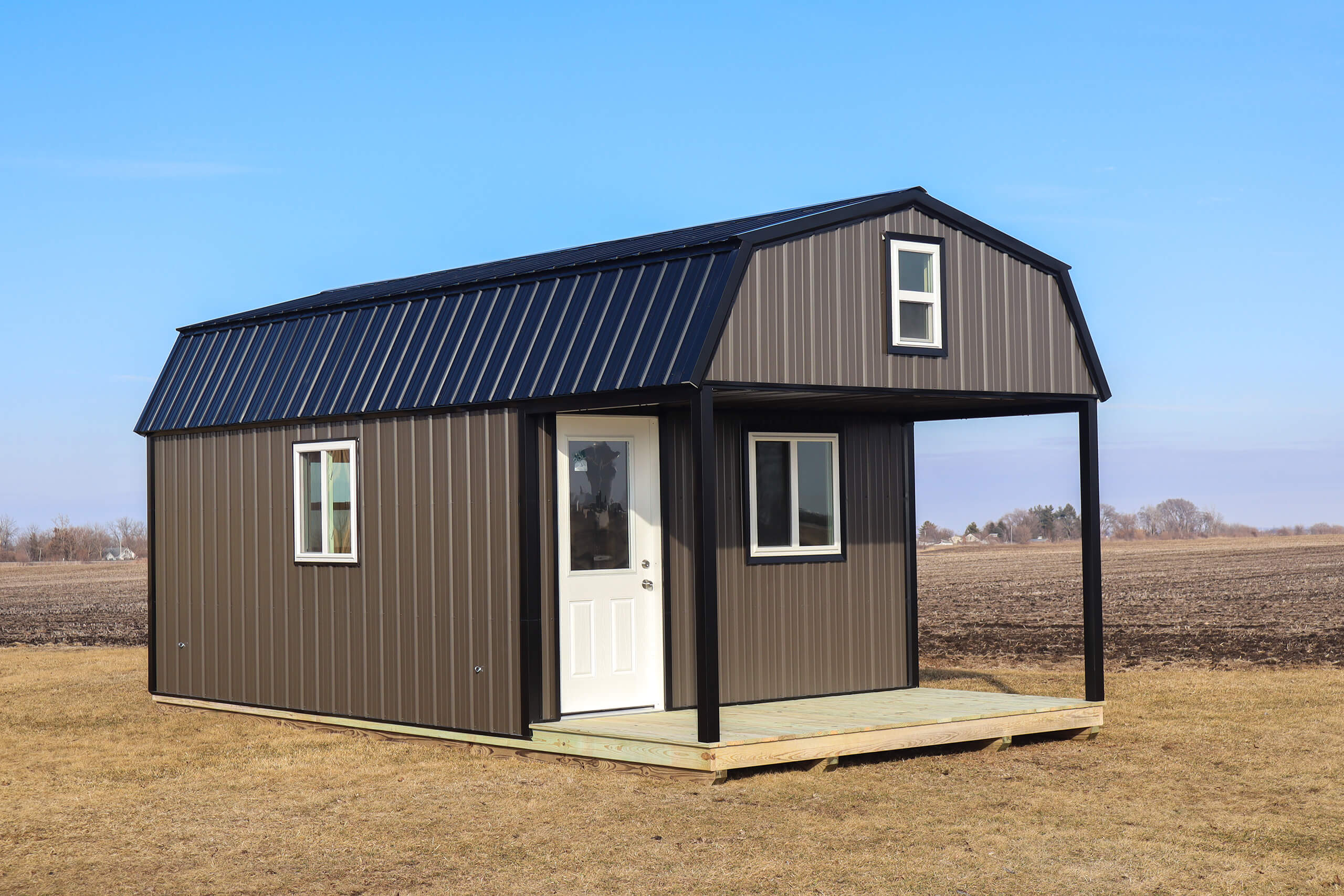A small, brown metal-sided shed with a white door and windows sits on a wooden deck in an open, grassy field under a clear blue sky.