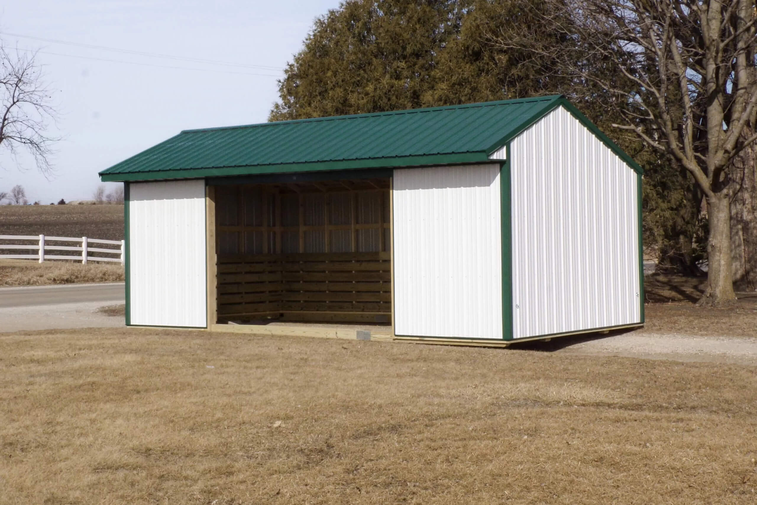 A white metal shed with a green roof and open front stands on dry grass near a road, surrounded by trees and a white fence.