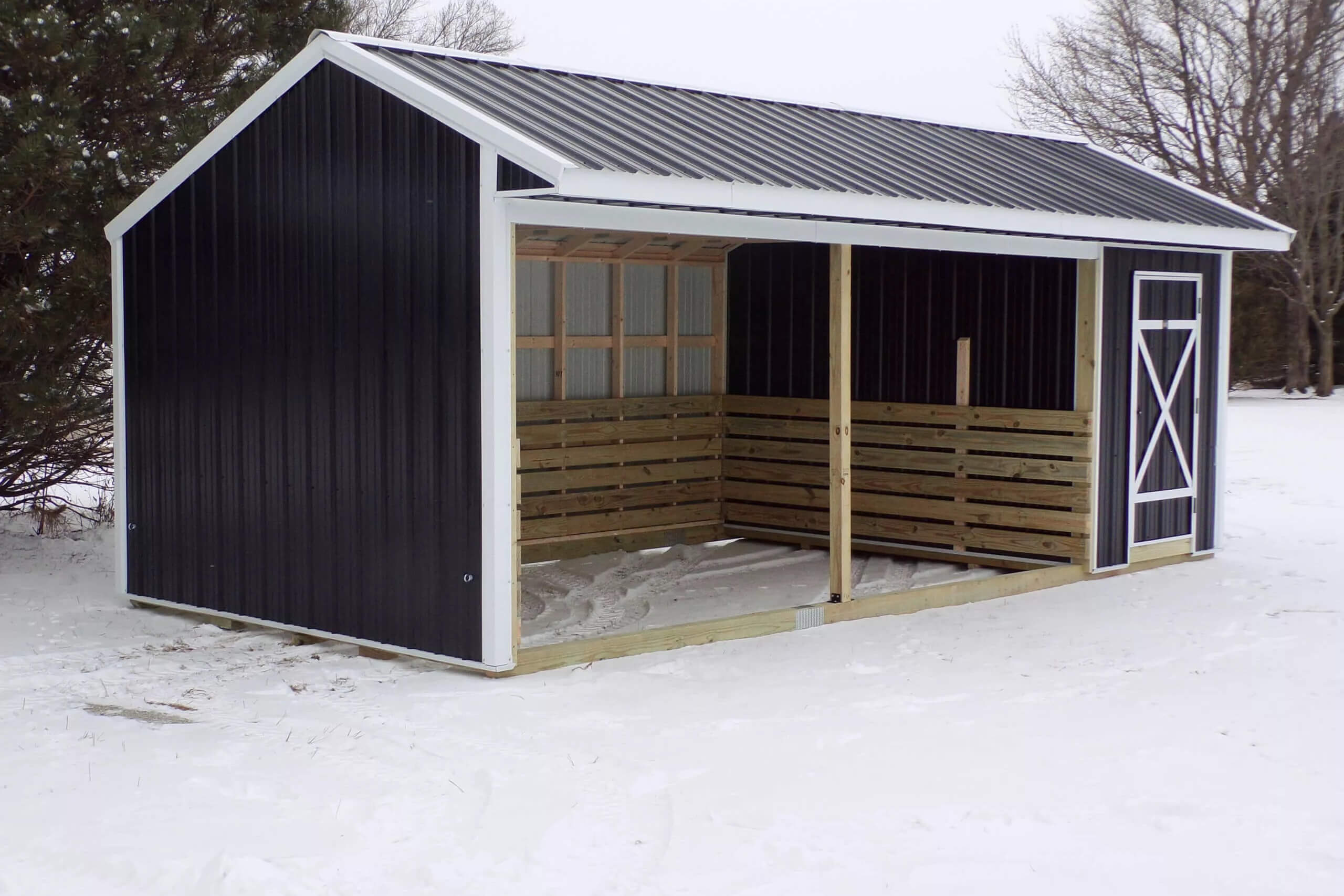 Black and white open-sided shed with a slanted metal roof, wooden slats, and a single door, standing on snowy ground with bare trees in the background.
