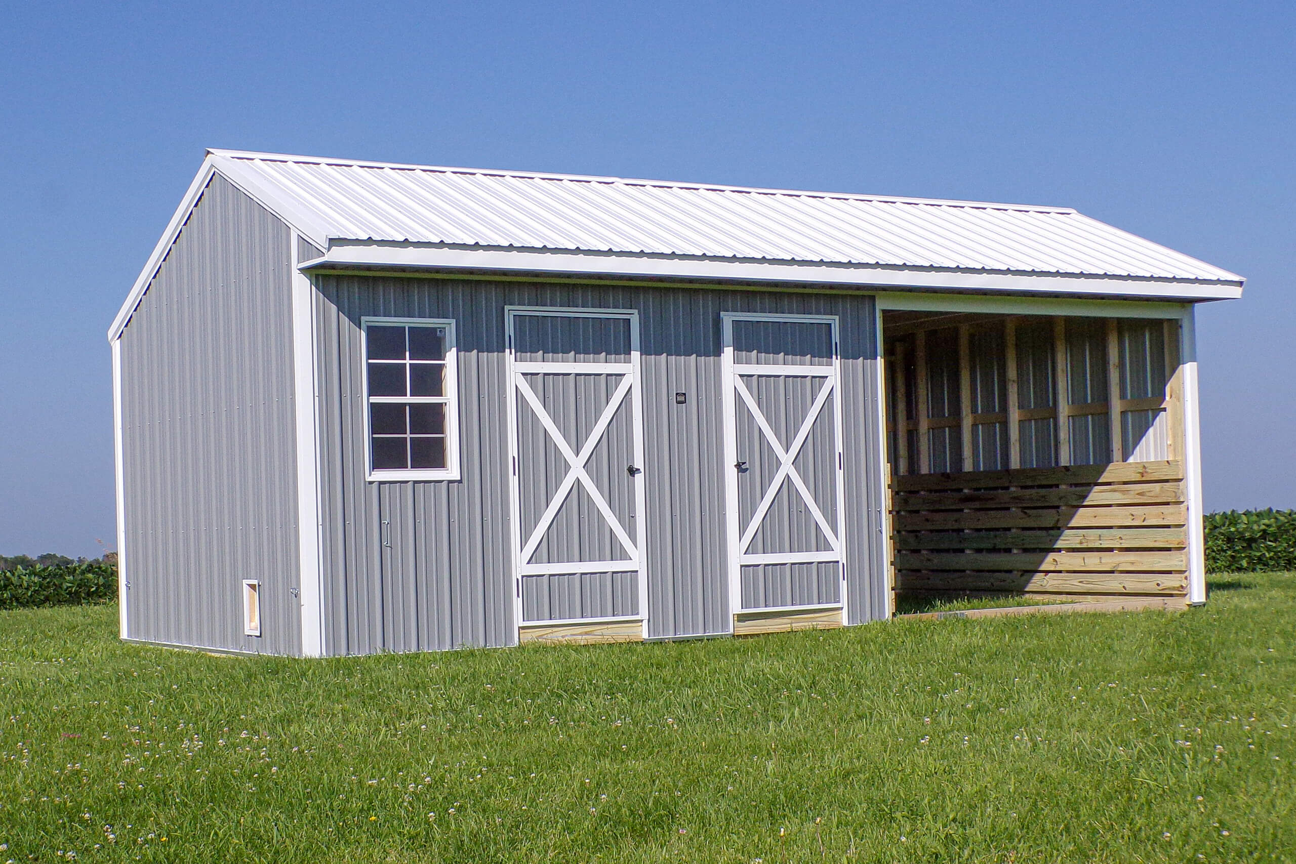 Gray metal storage shed with two doors, a window, and an attached open-sided section, situated on green grass under a clear blue sky.