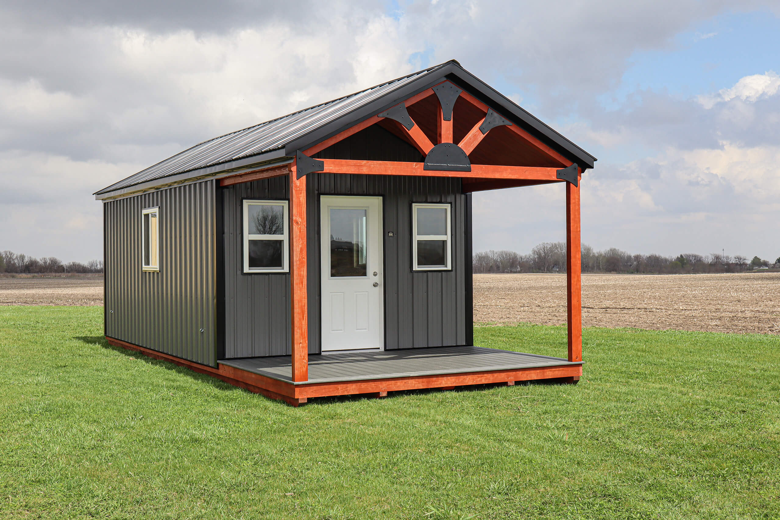 Small dark gray cabin with a white door, two windows, and a covered porch, sitting on a grassy field under a partly cloudy sky.