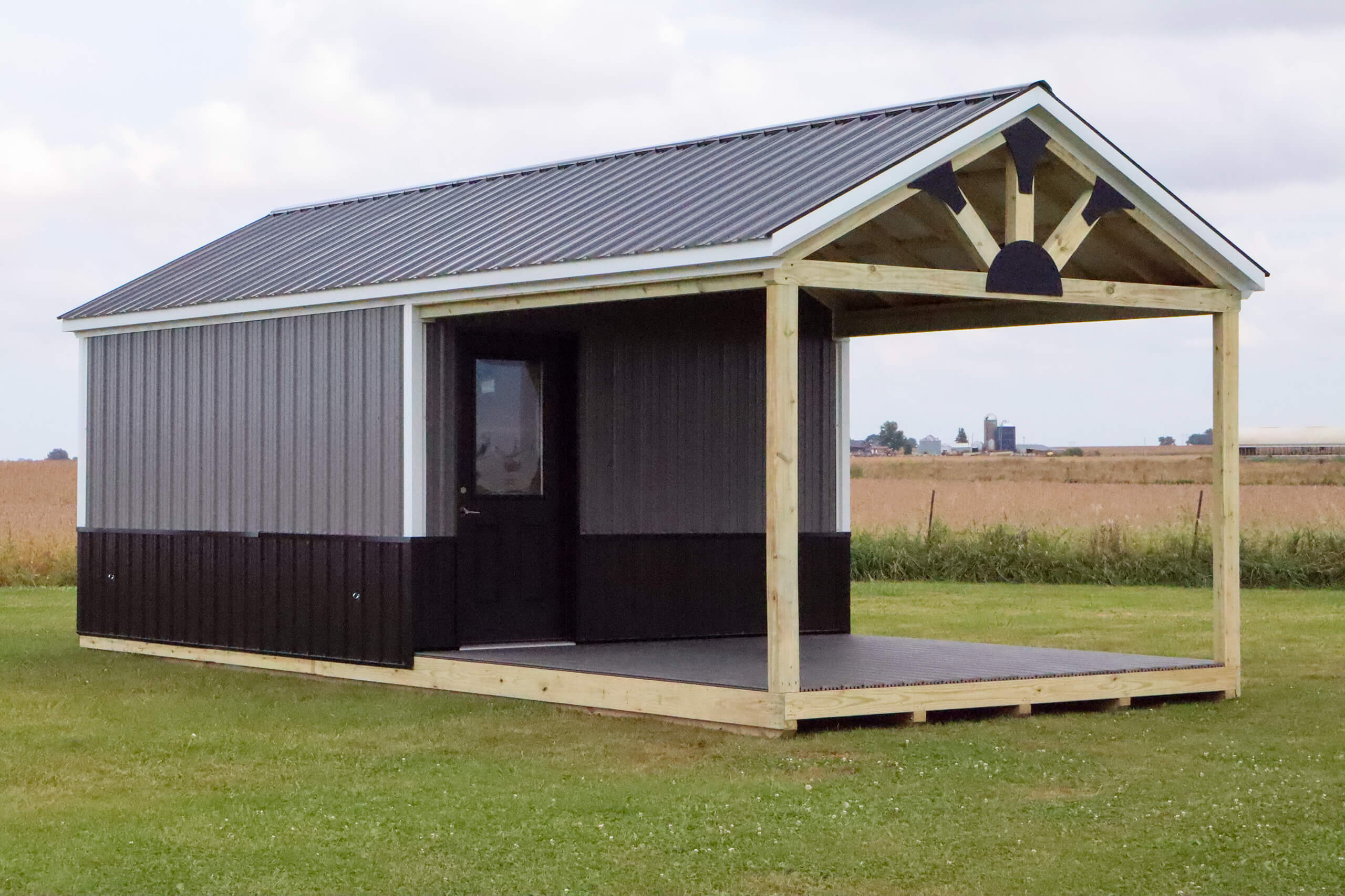 A small, gray and black shed with a covered porch stands on a grassy field. The shed has a metal roof and a single door.