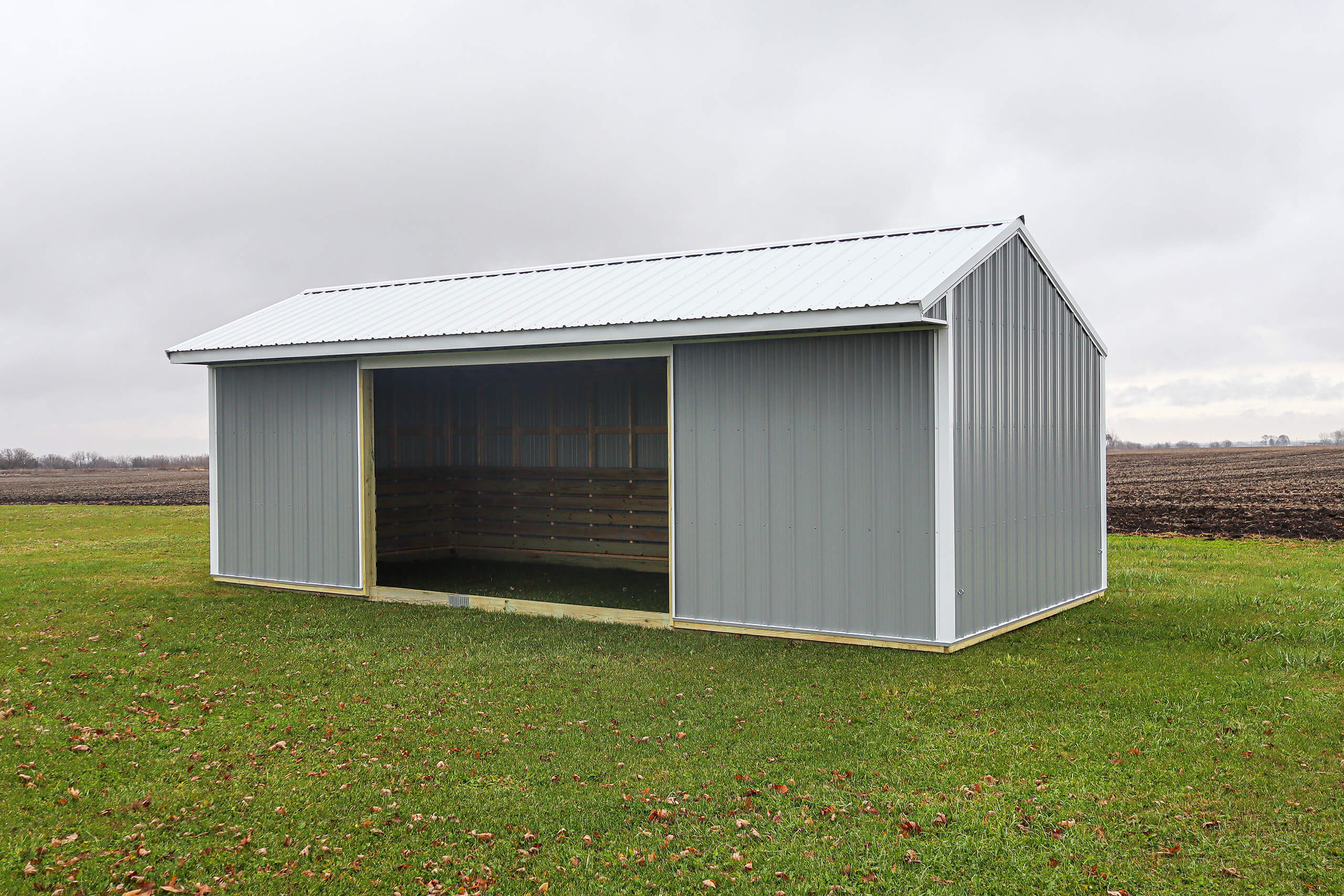 A gray metal shed with an open front stands on a grassy field near tilled farmland under a cloudy sky.
