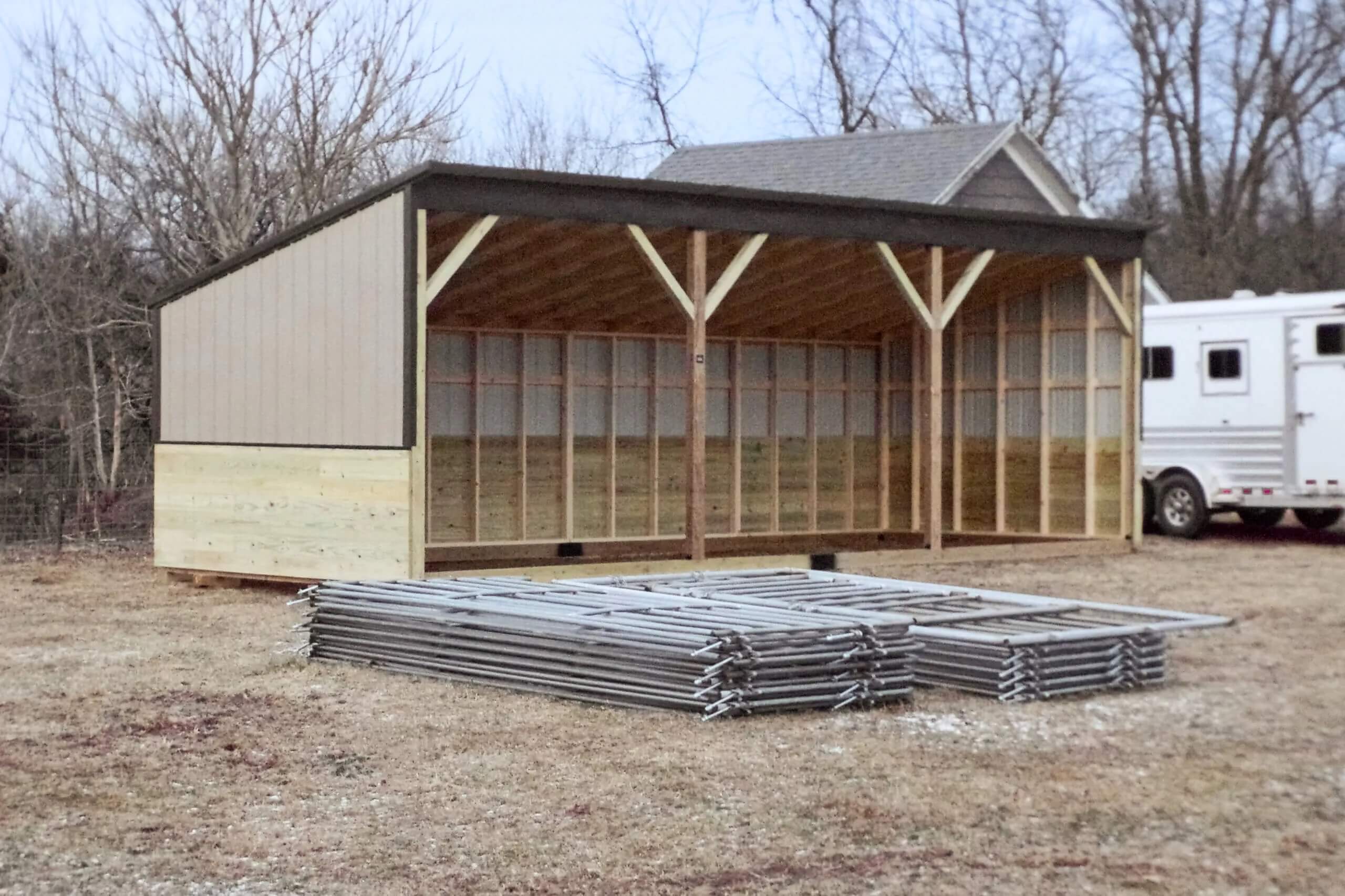 A partially finished open shed with exposed framing sits behind stacks of metal fencing panels on a dry, grassy lot. Trees and a white trailer are in the background.