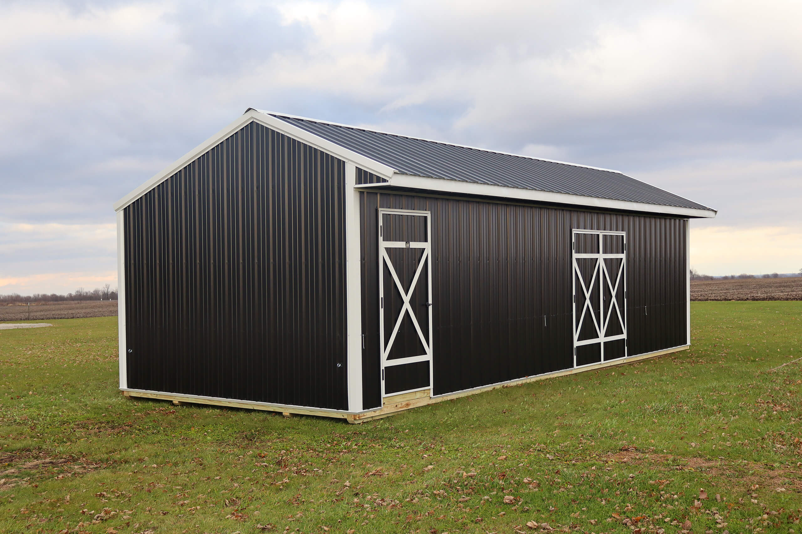 A black metal barn with white trim and double doors stands on a grassy field under a cloudy sky.