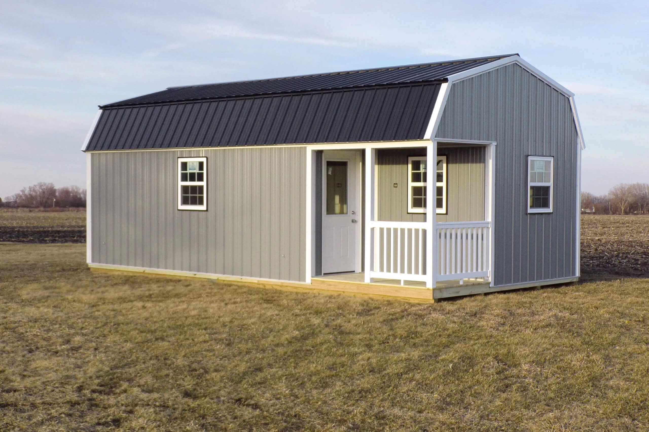Small gray shed with white trim and a black metal roof, featuring a door, two windows, and a small covered porch, situated on a grassy field.