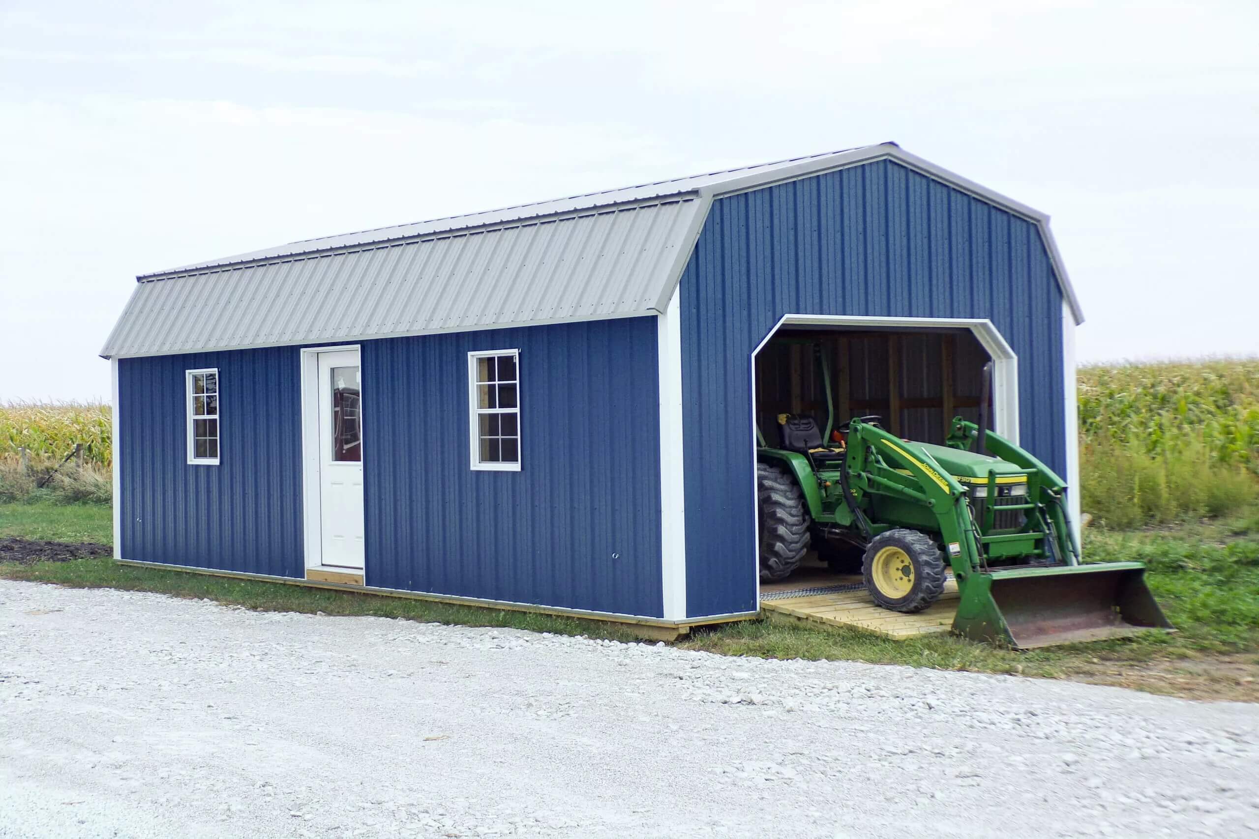 A green tractor is parked inside a blue metal shed with a white door and windows, situated next to a gravel path.