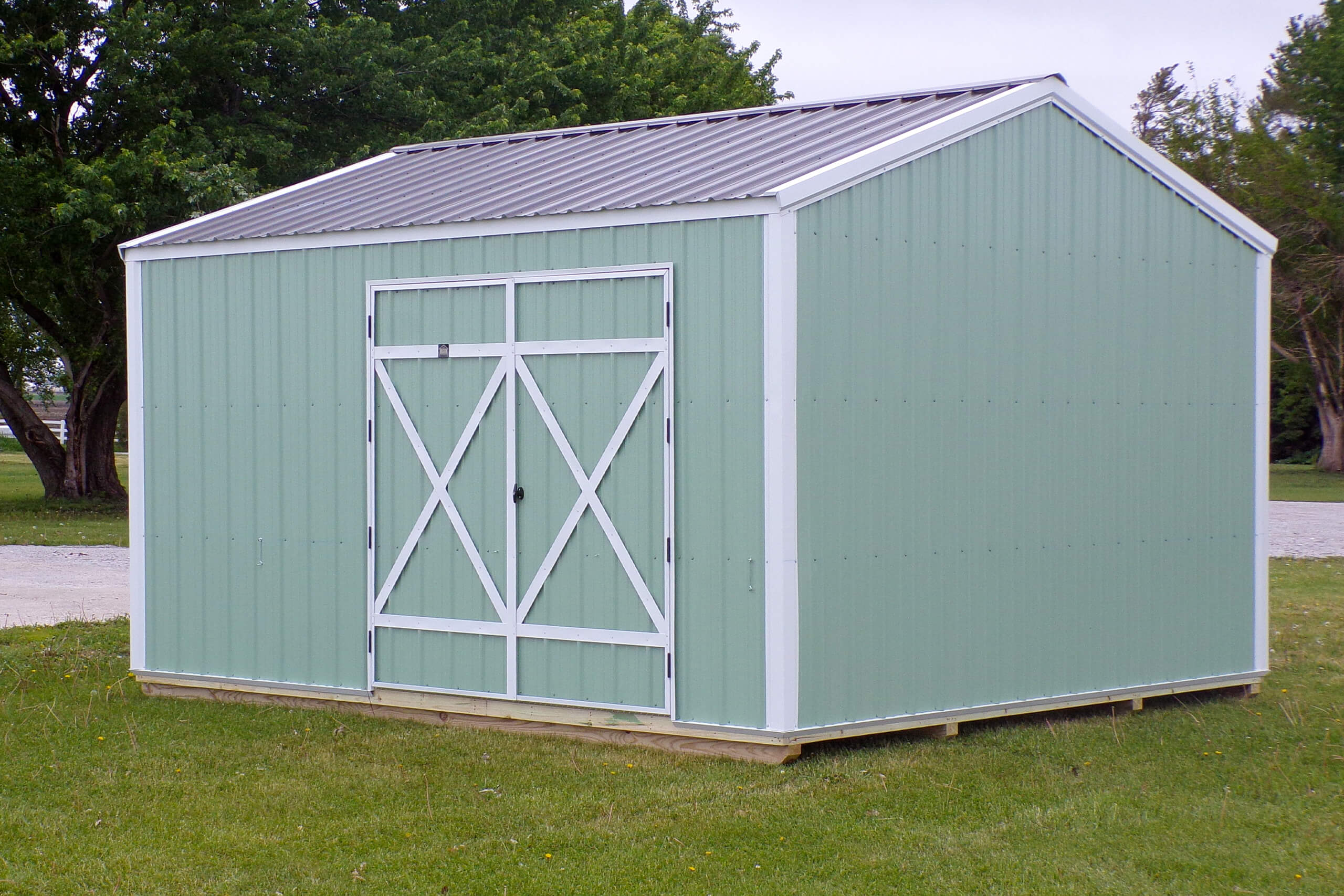 Light green metal storage shed with a gable roof and double doors, standing on a grassy lawn with trees in the background.