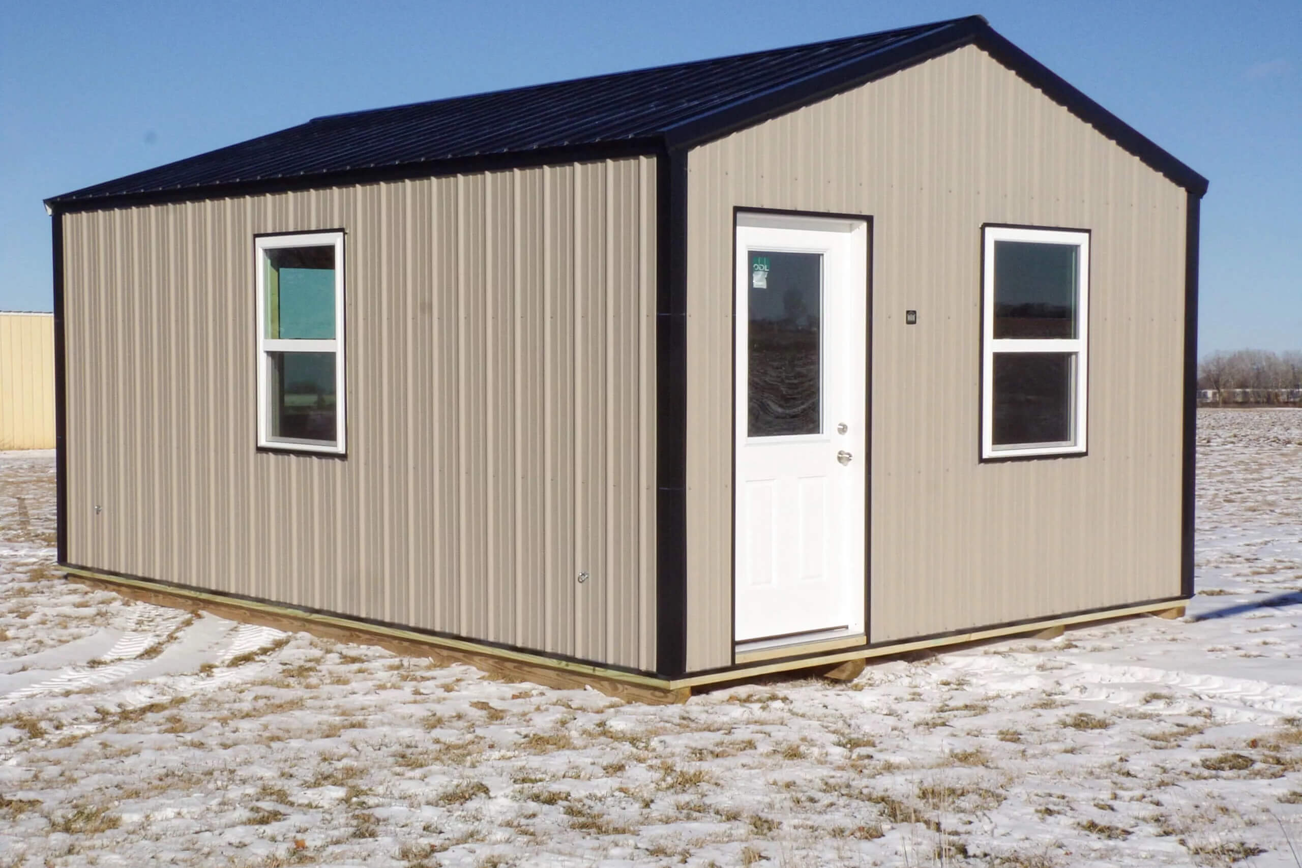 Small, single-story metal building with a black roof, two windows, and a white door, set on a snowy, grassy field under a clear blue sky.