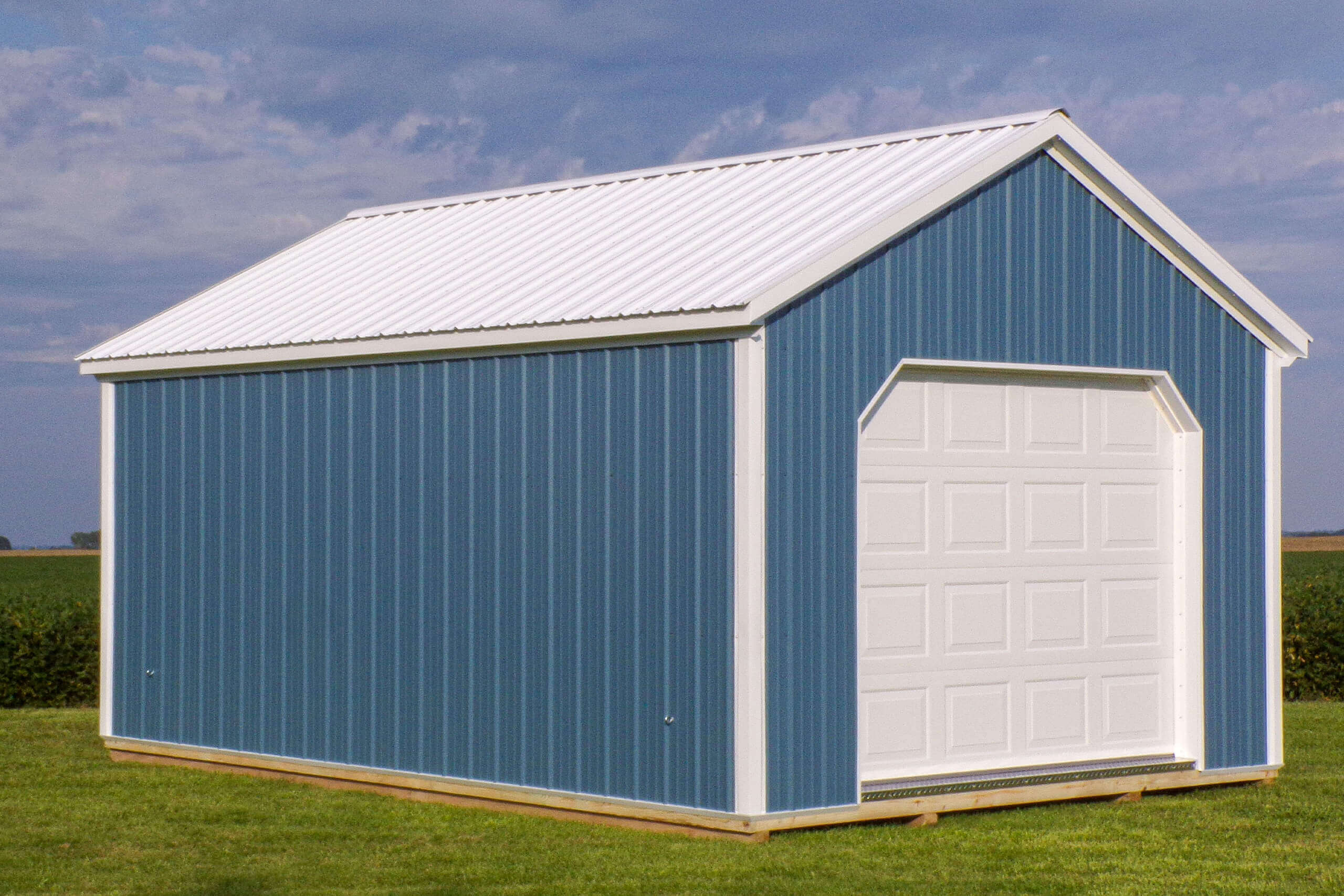 A blue metal shed with a white roof and garage door sits on a grassy area under a partly cloudy sky.
